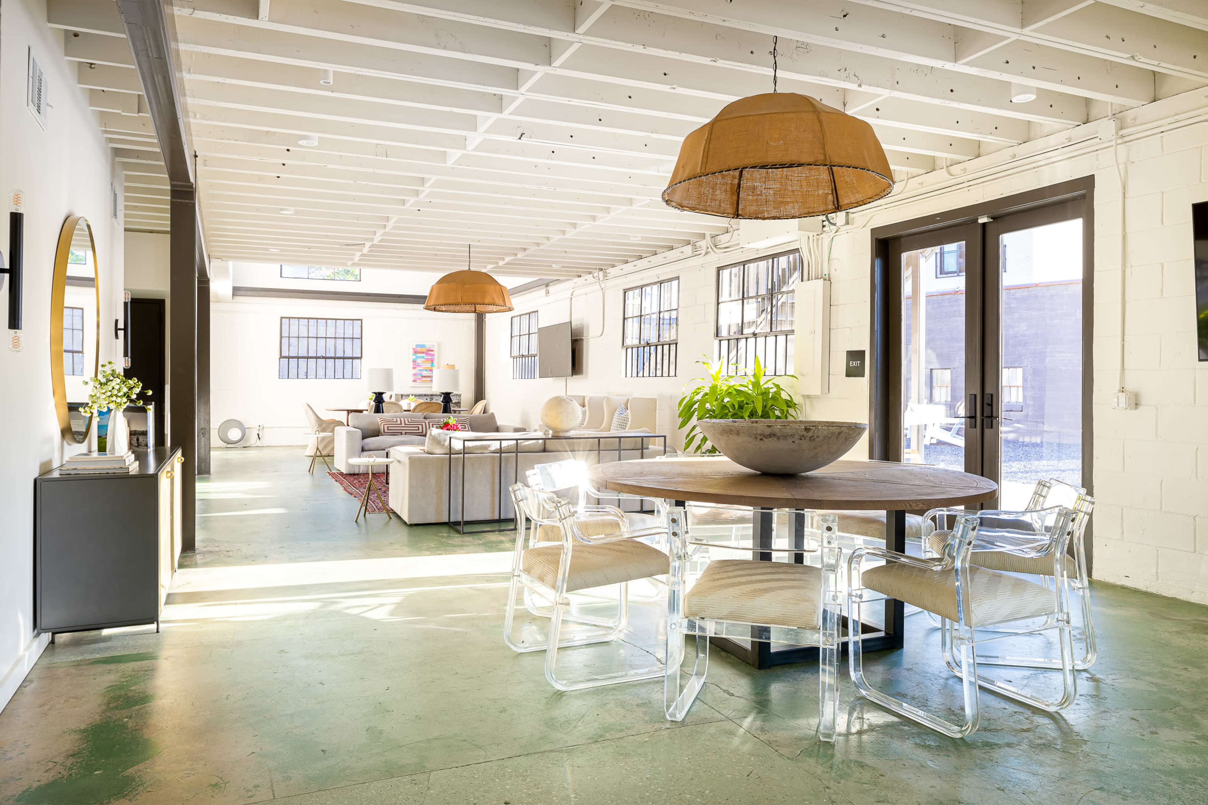 A modern interior space with a round dining table surrounded by clear chairs, adjacent to a living area with a sofa and large windows.