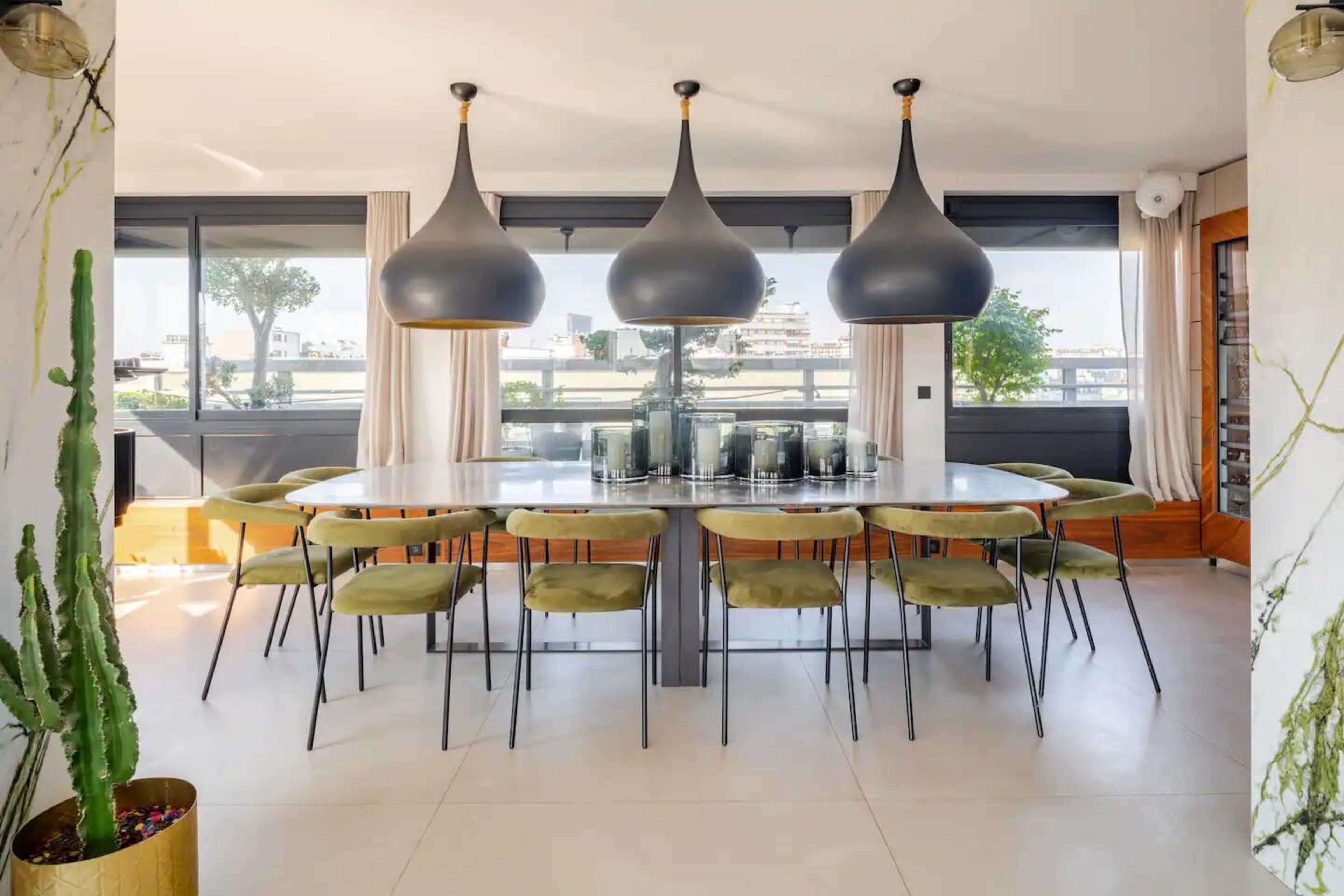 A modern dining room features a large table surrounded by green chairs, with three pendant lights hanging above and large windows providing a view outside.