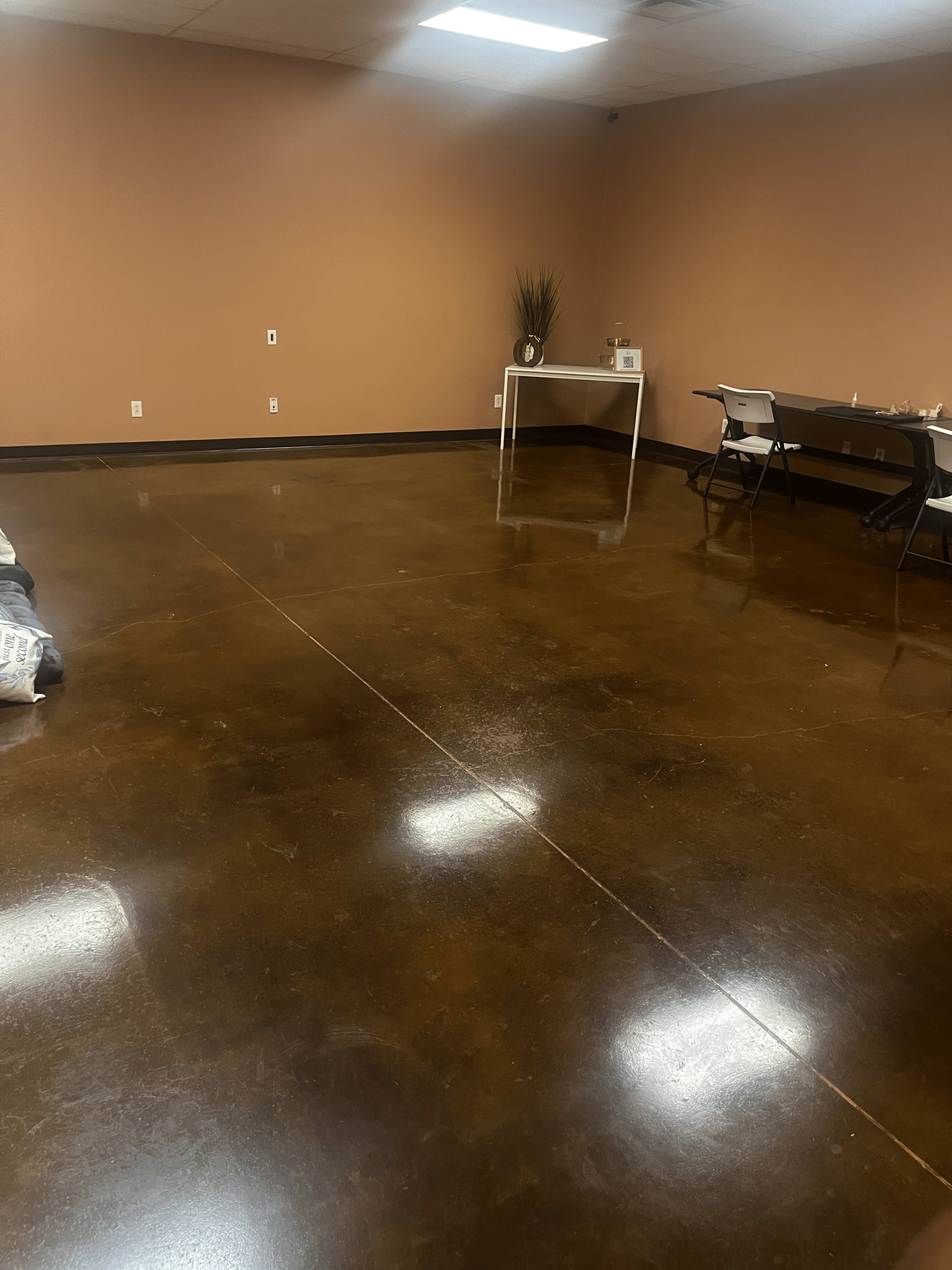 A large, empty room with a polished brown concrete floor, a small table, and two chairs against a wall.