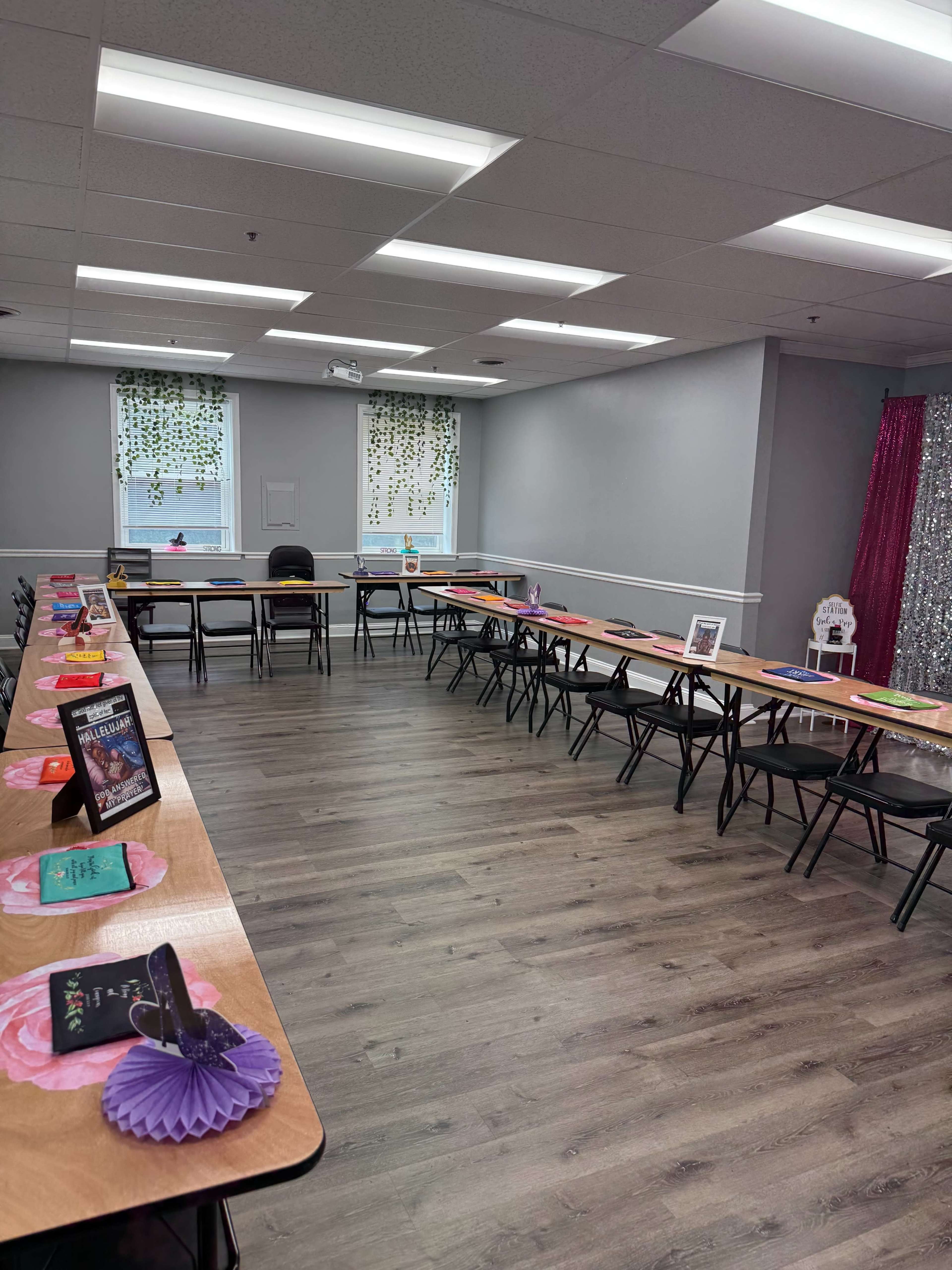 The image shows a spacious room arranged with long tables set in a U-shape, decorated with colorful items and surrounded by chairs.