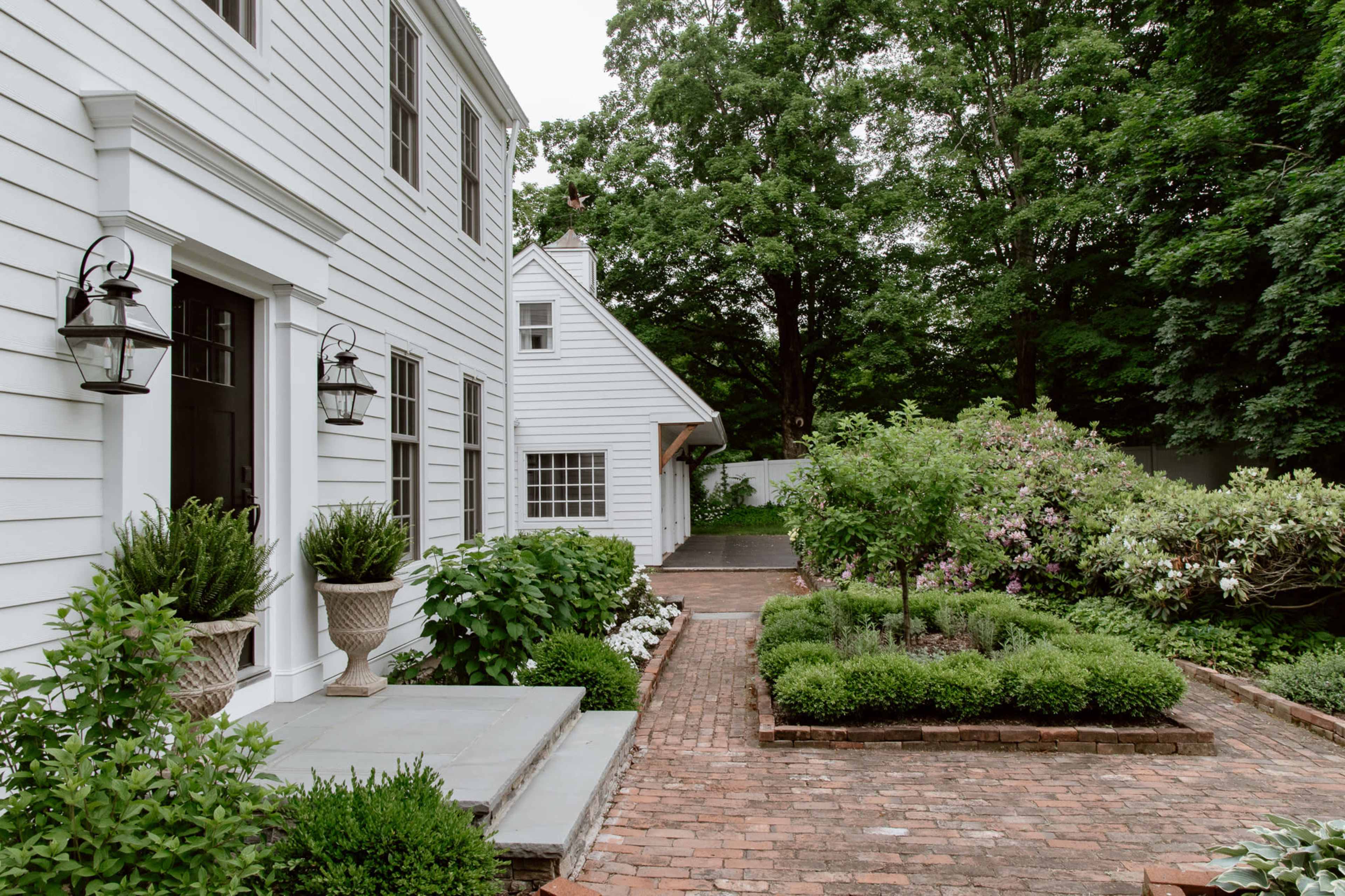 A white-sided house features a brick walkway bordered by lush greenery and flowering plants.