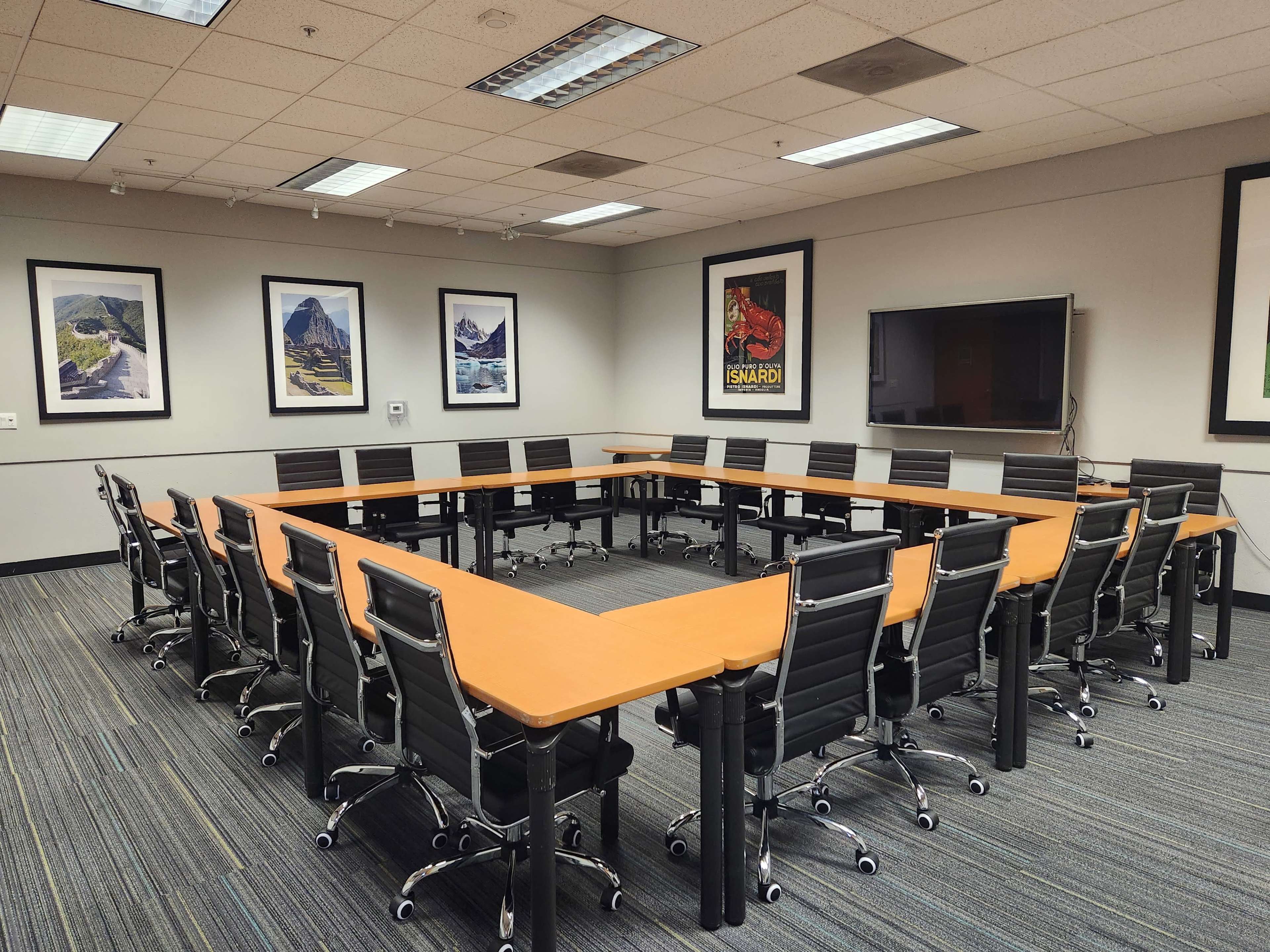 The image shows a spacious conference room featuring a large rectangular wooden table surrounded by black office chairs, with framed landscape posters on the walls and a television mounted in the corner.