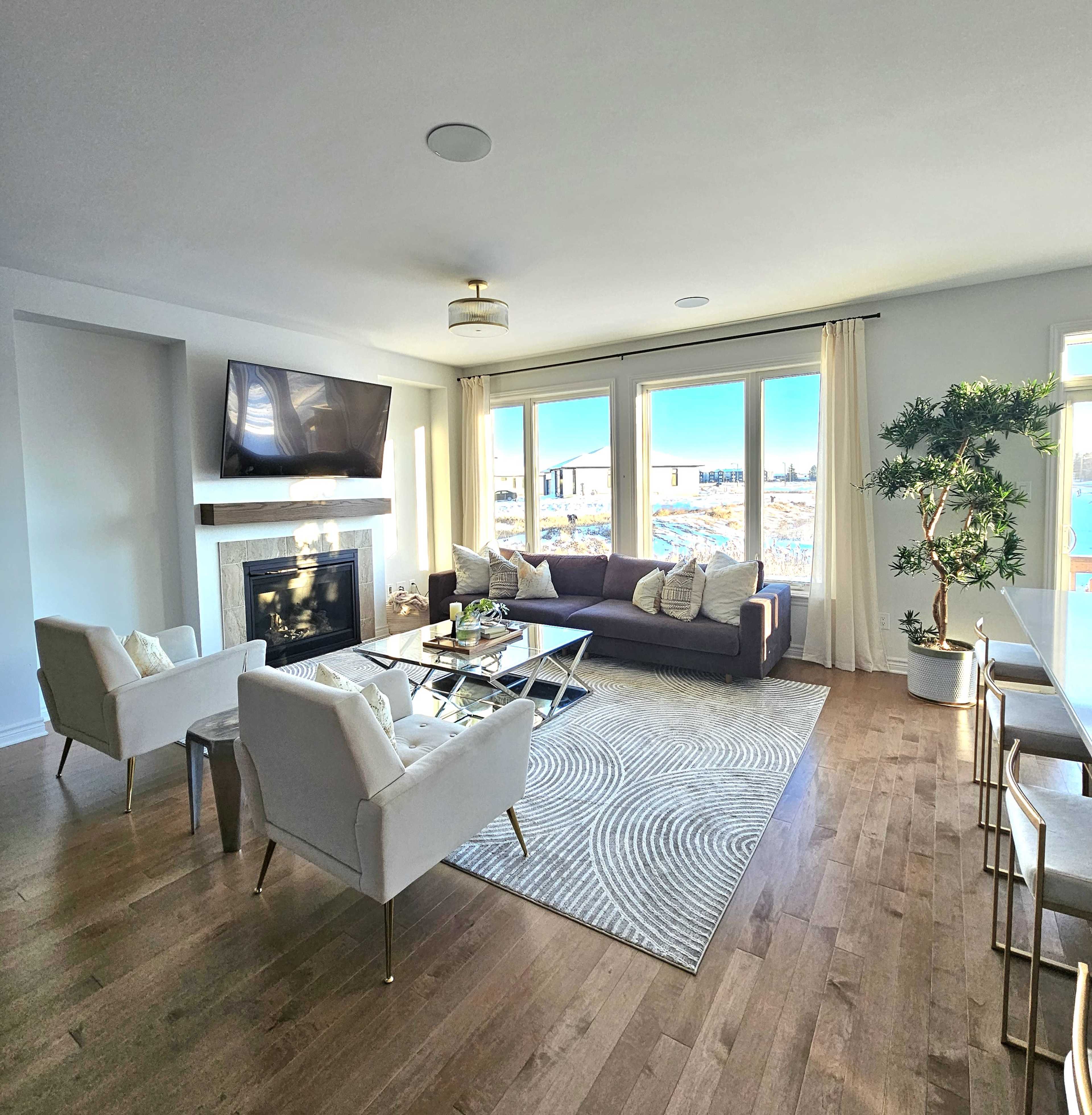 A modern living room features a dark sectional sofa, two white armchairs, a glass coffee table, and large windows overlooking a snowy landscape.