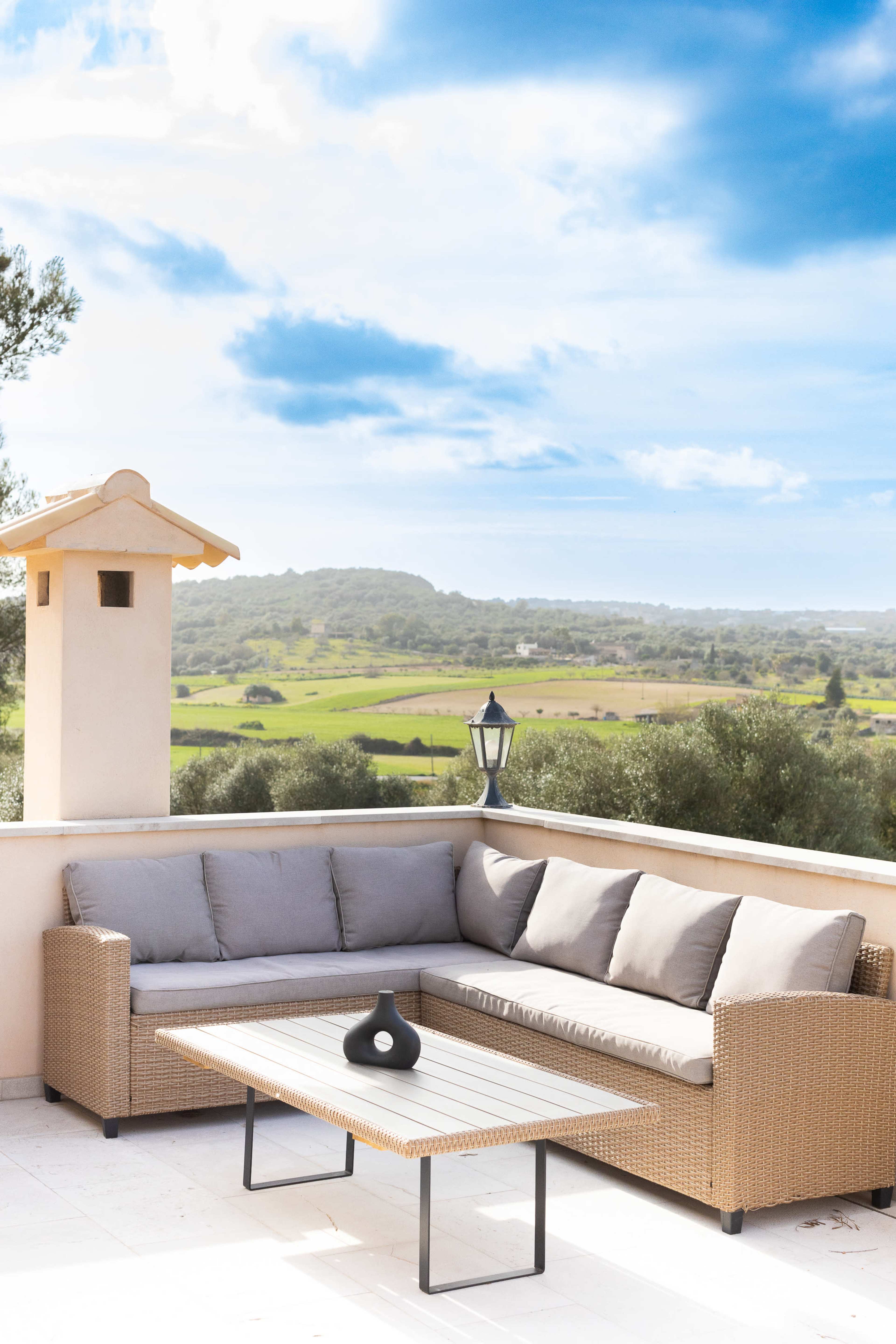 A corner outdoor seating area with a gray cushioned sofa and a low table overlooks a green landscape under a partly cloudy sky.