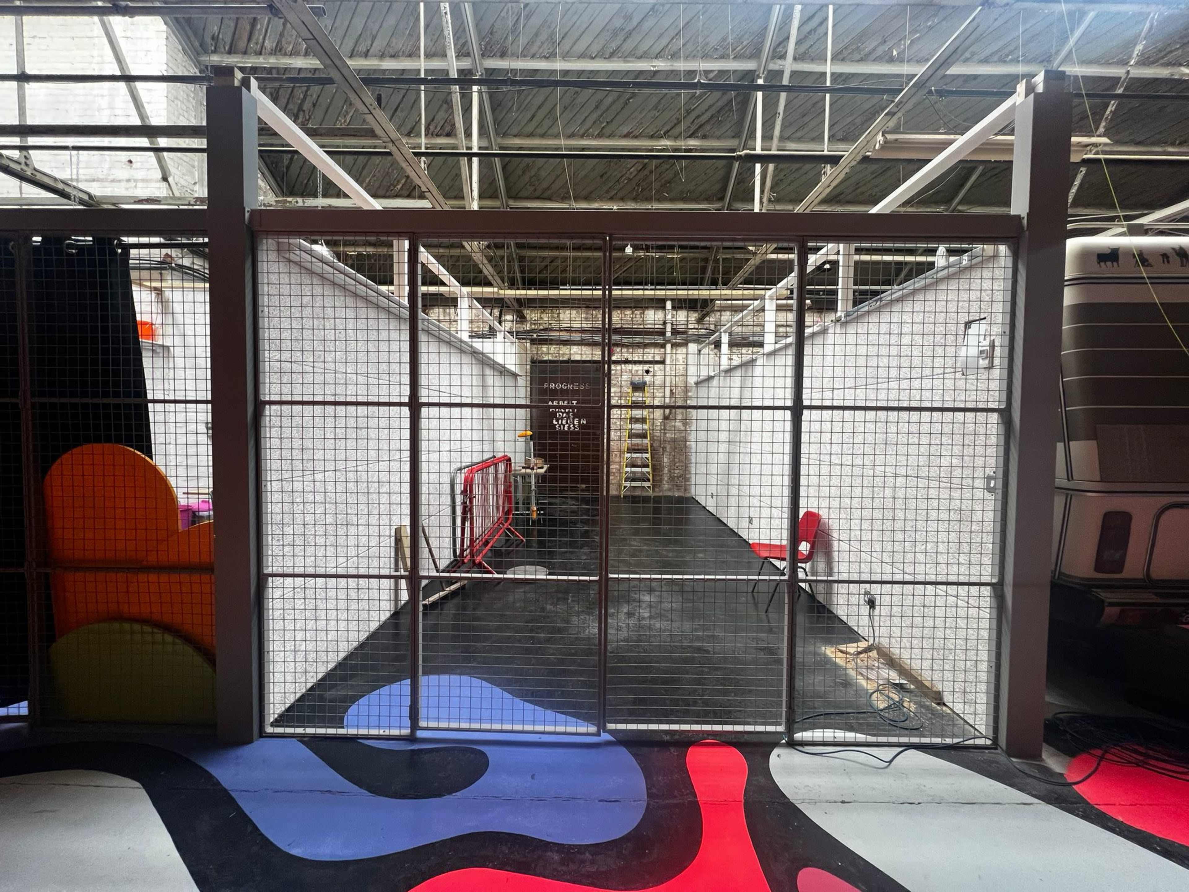 An empty industrial space features a gated entrance that leads to a long hallway with a wooden ladder against a wall in the background.
