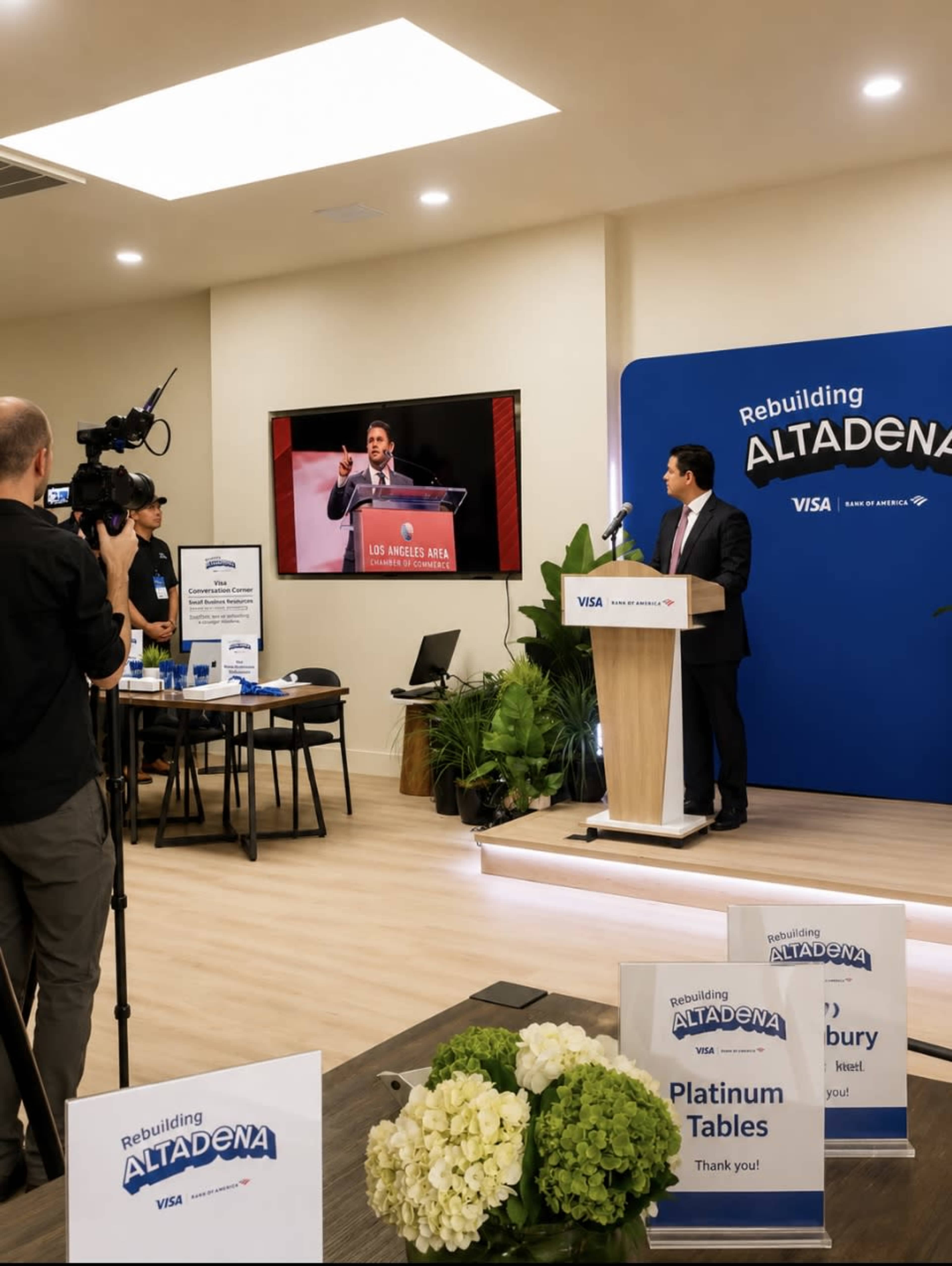 A man speaks at a podium during an event titled "Rebuilding Altadena," while a camera operator records and a screen displays a presenter.