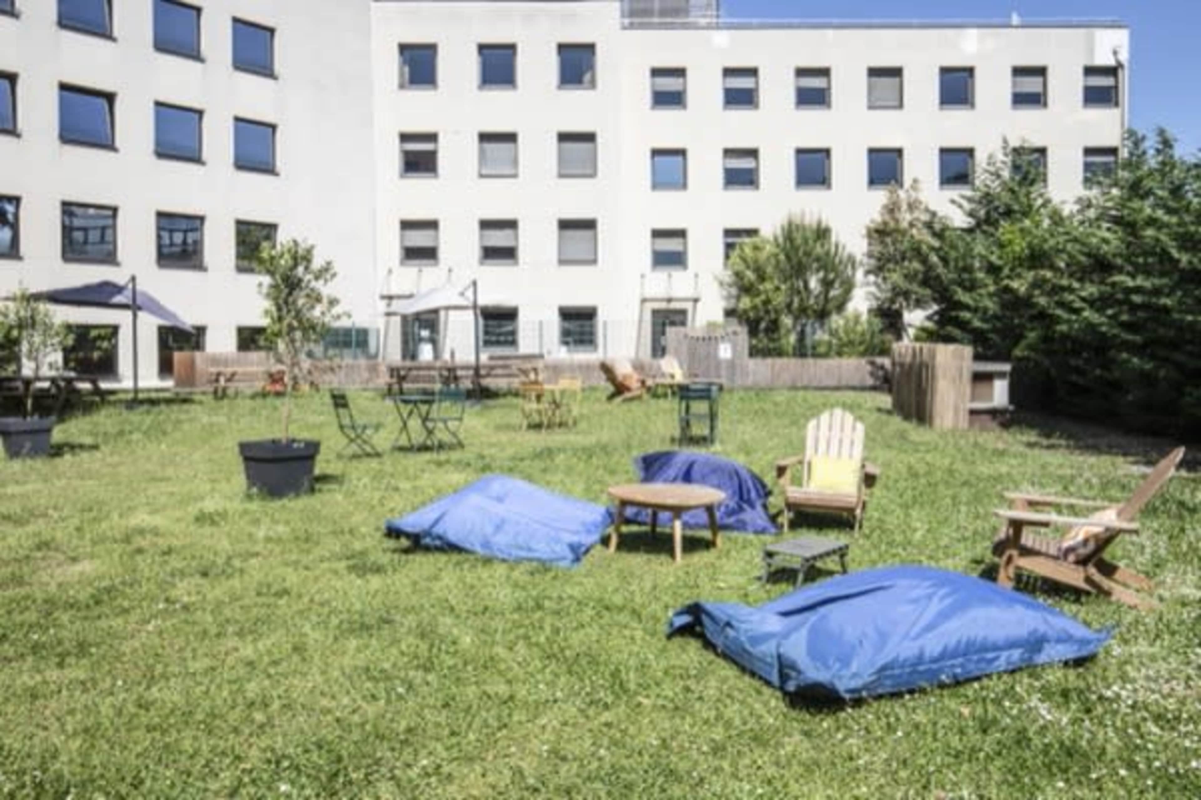 An outdoor area with blue cushions, wooden chairs, and tables on a grassy lawn surrounded by a building with multiple windows.