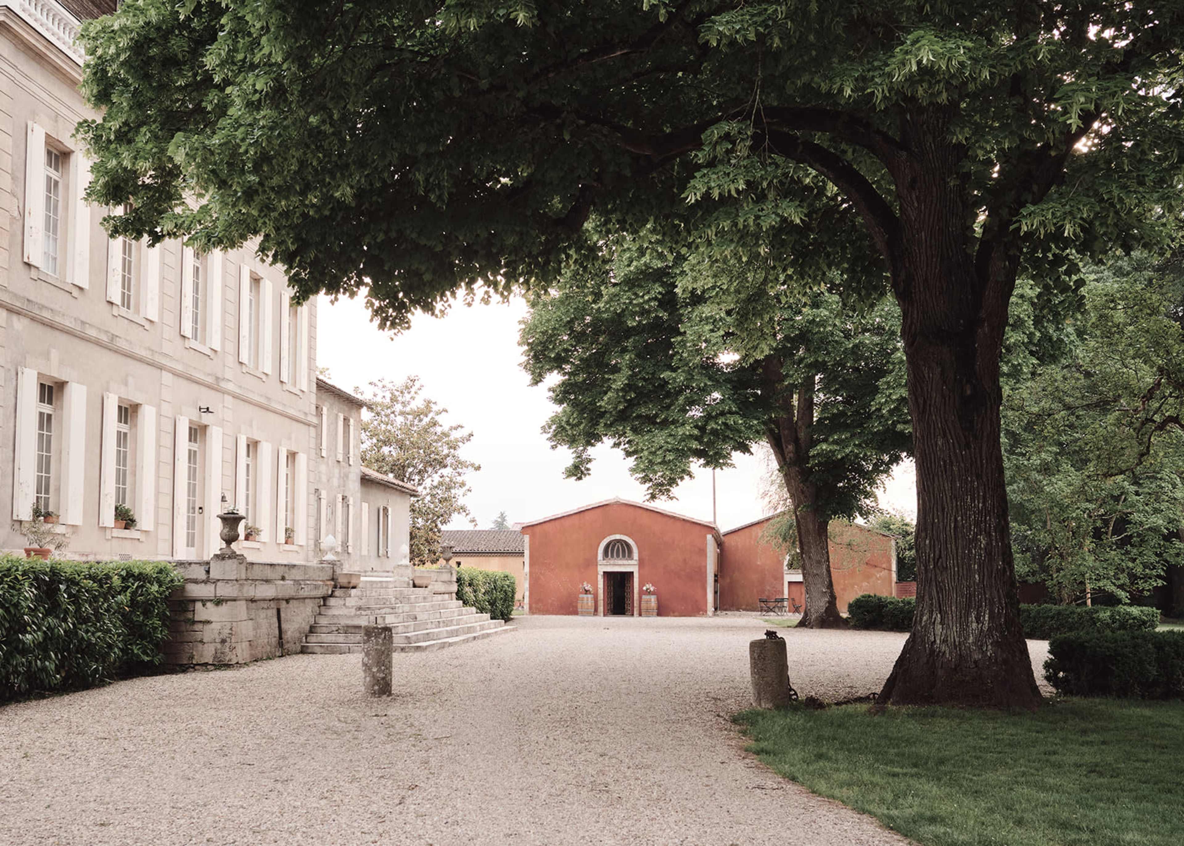 A gravel pathway leads to a large house on the left, while a red building stands in the background, framed by a large tree.