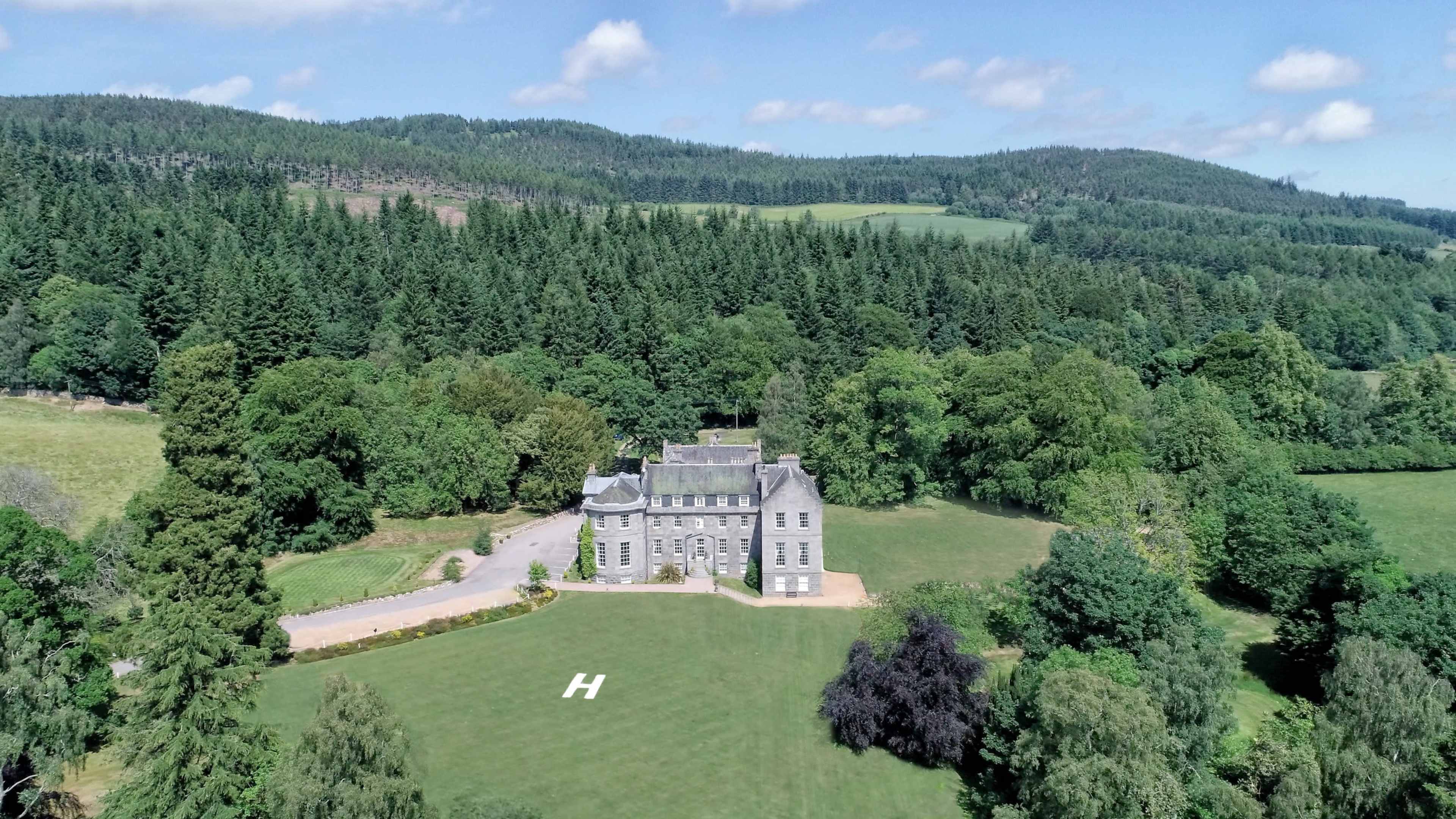 A large, historic mansion is surrounded by lush greenery and rolling hills, with a helicopter landing pad marked by an "H" on the lawn.