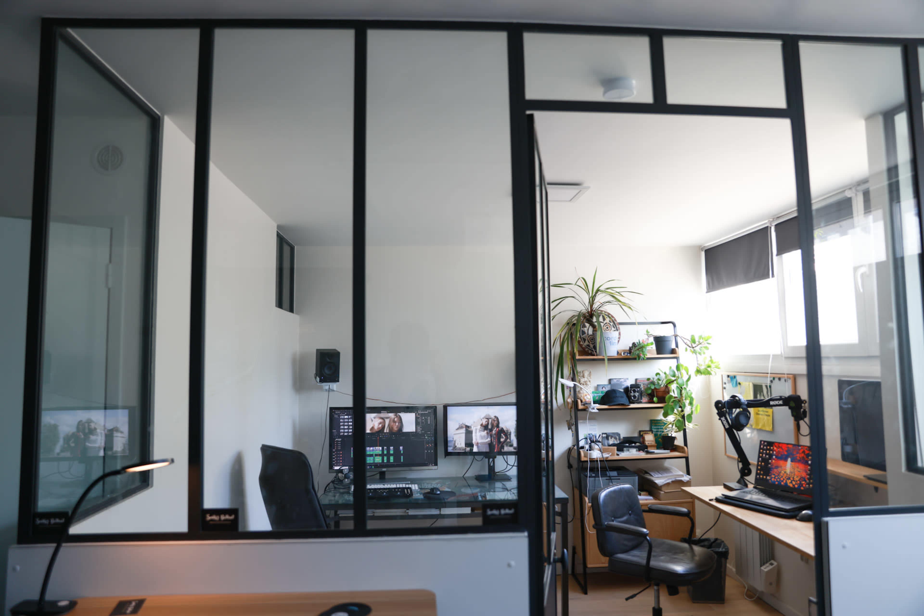 The image shows a modern office space divided by glass walls, featuring two separate work areas with computers, office furniture, and shelves filled with plants and equipment.