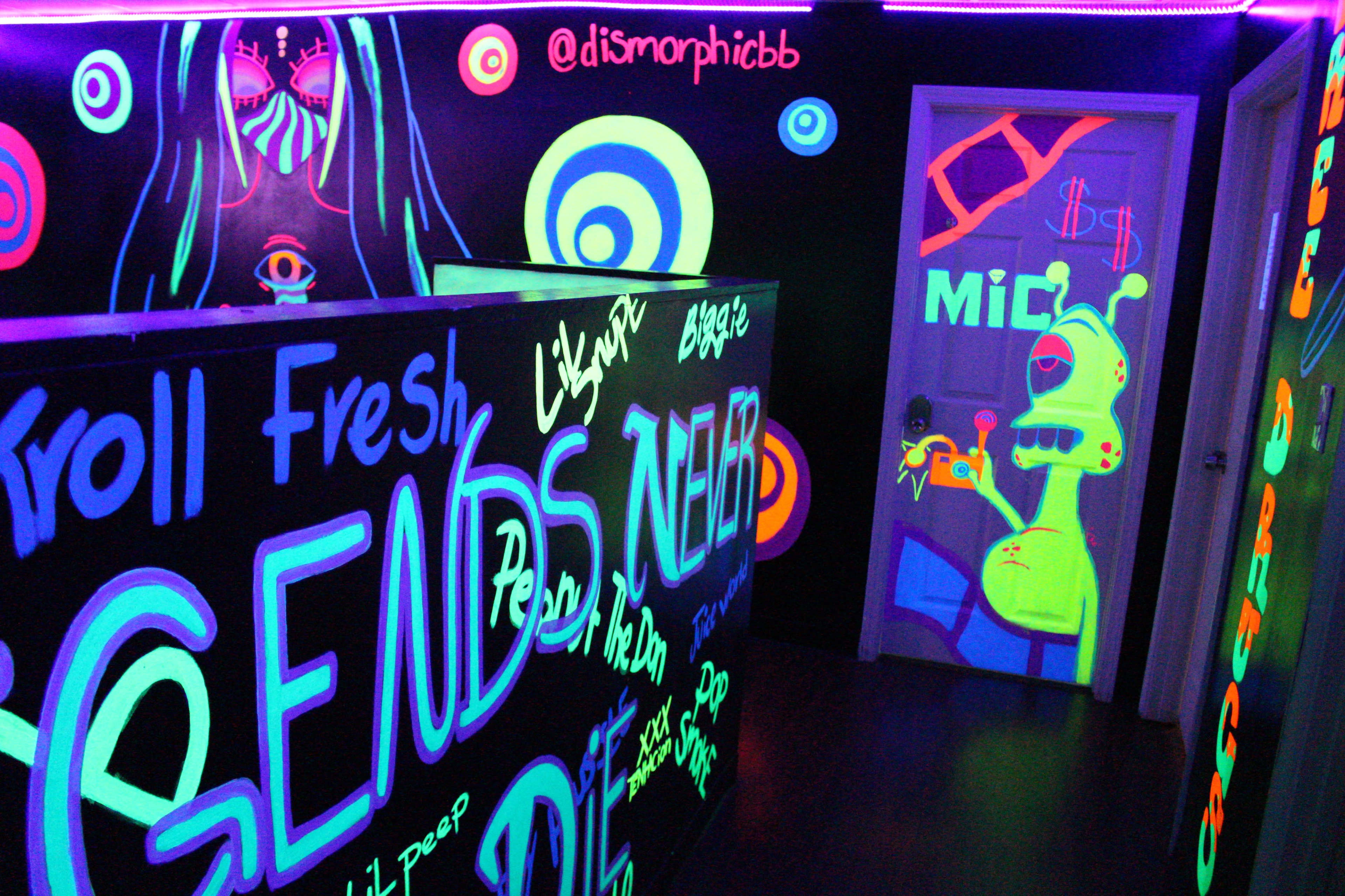 The image shows a room decorated with fluorescent graffiti and designs, including abstract shapes and text, illuminated under black lights.