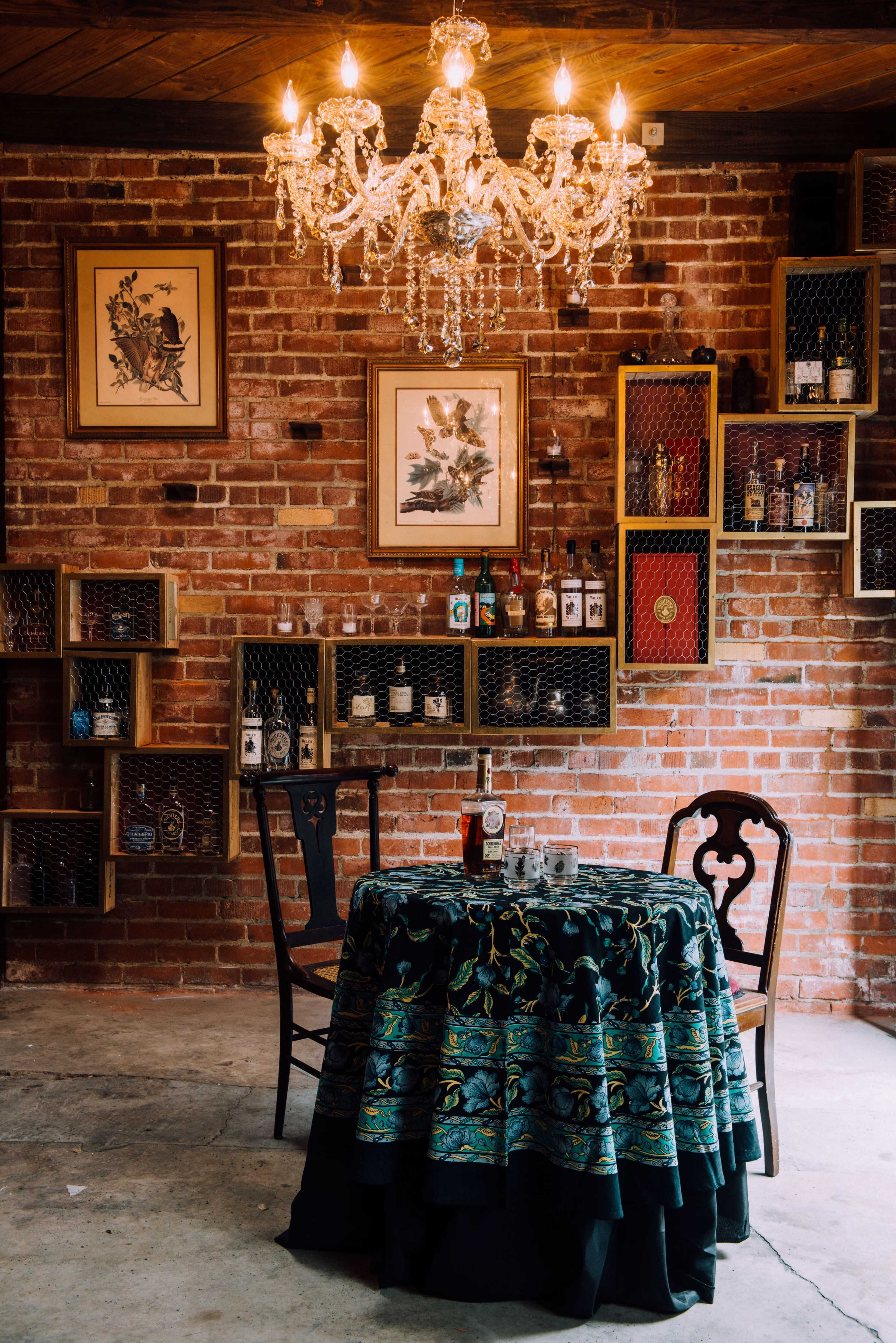 A decorative table with a patterned cloth is set in a brick-walled room adorned with a chandelier and shelves displaying various bottles.
