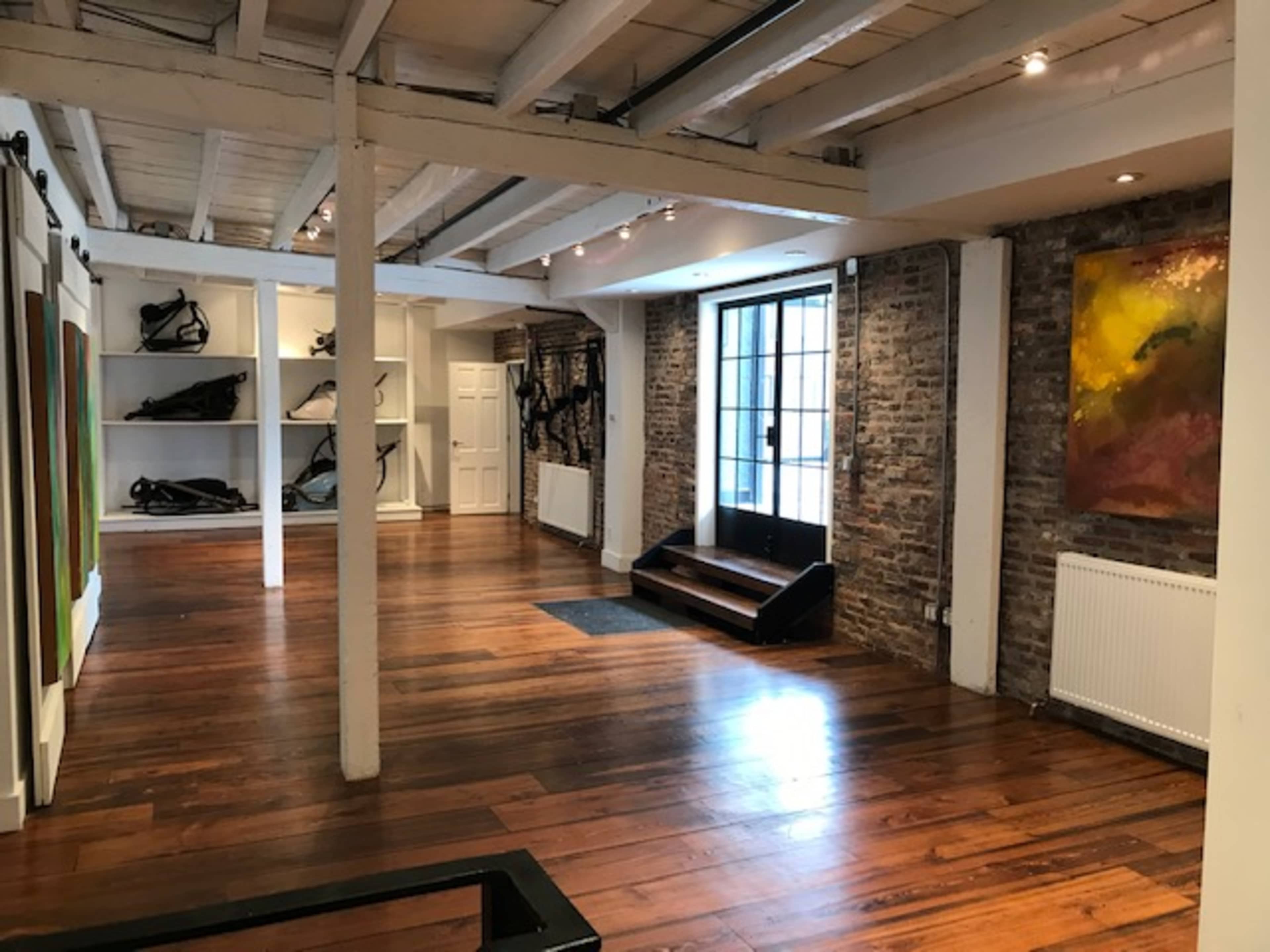 The image shows a spacious interior with wooden flooring, exposed brick walls, and a large window allowing light to enter.