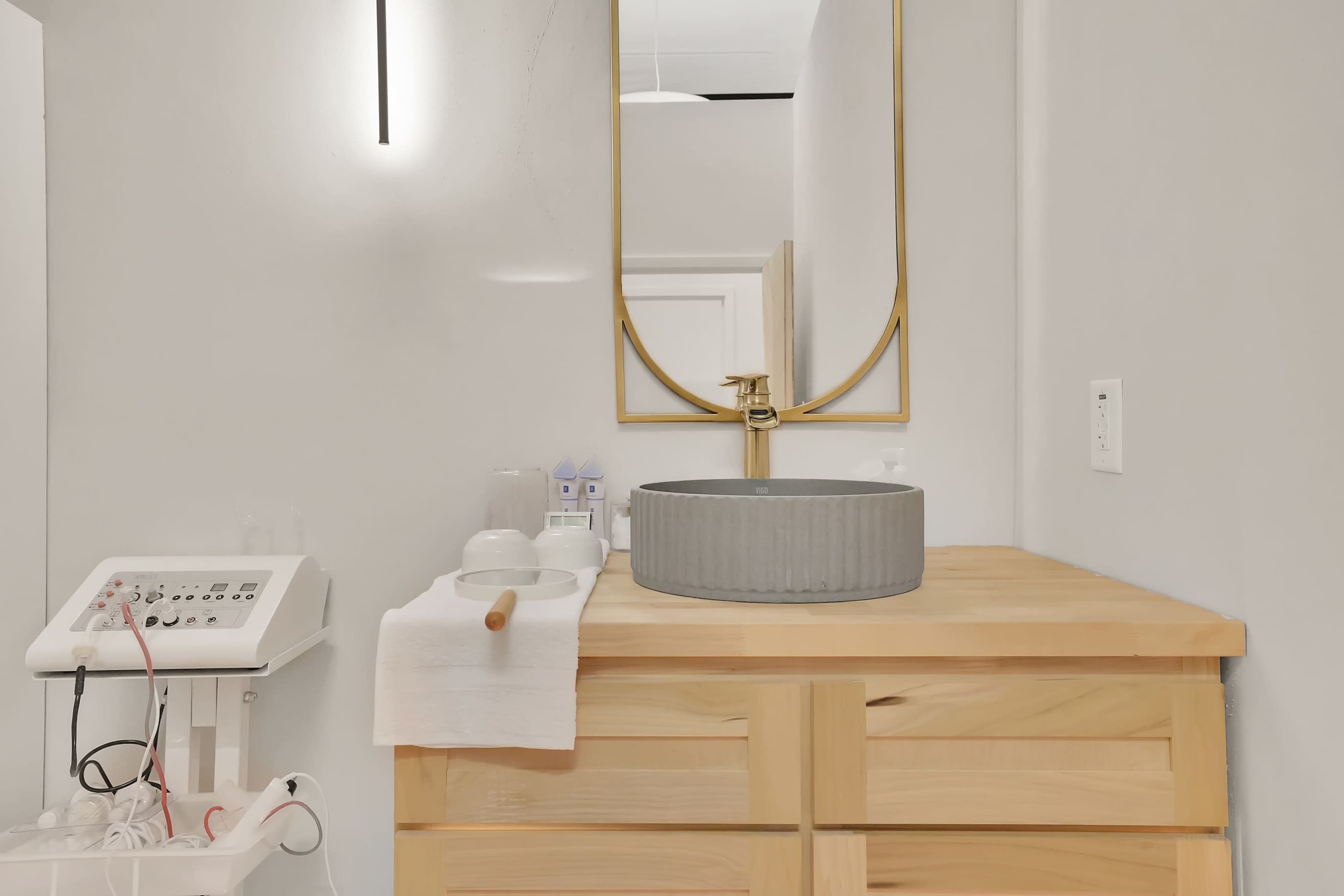 The image shows a modern bathroom vanity with a round stone sink, a wooden countertop, and a large mirror above it, accompanied by skincare products and a facial machine beside it.