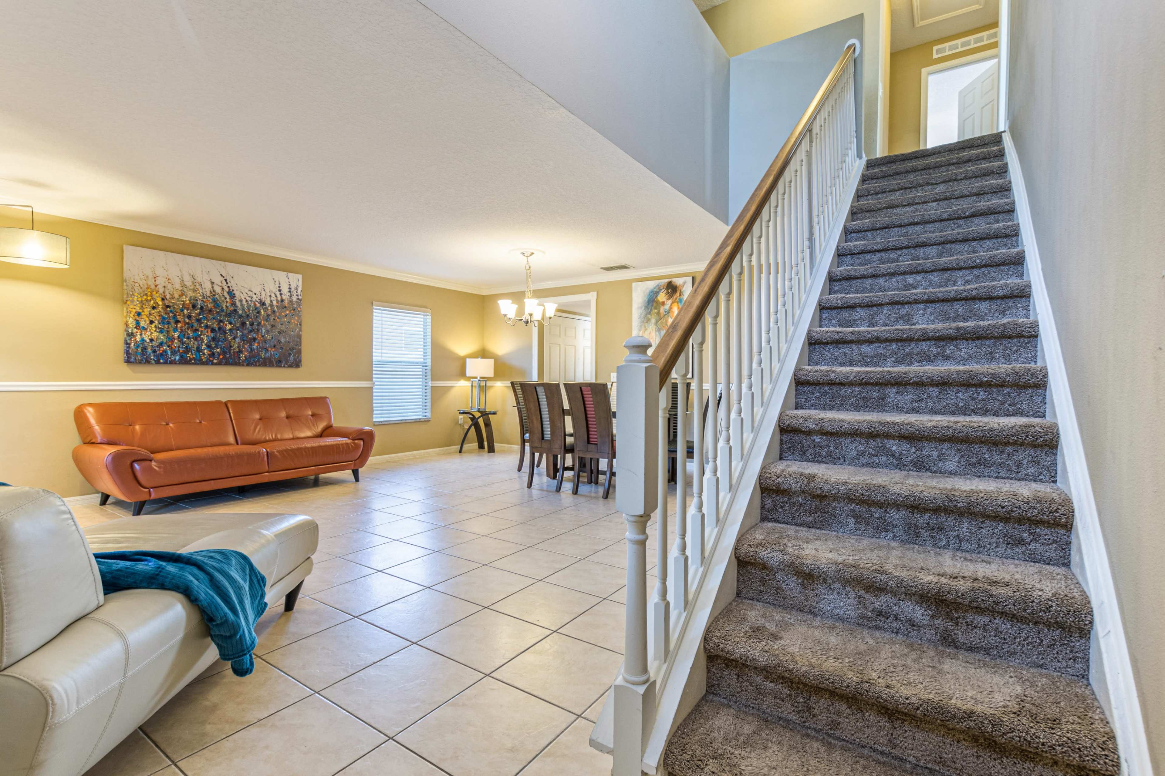 A living area with a staircase leading to an upper level, featuring a couch, a dining table, and artwork on the walls.