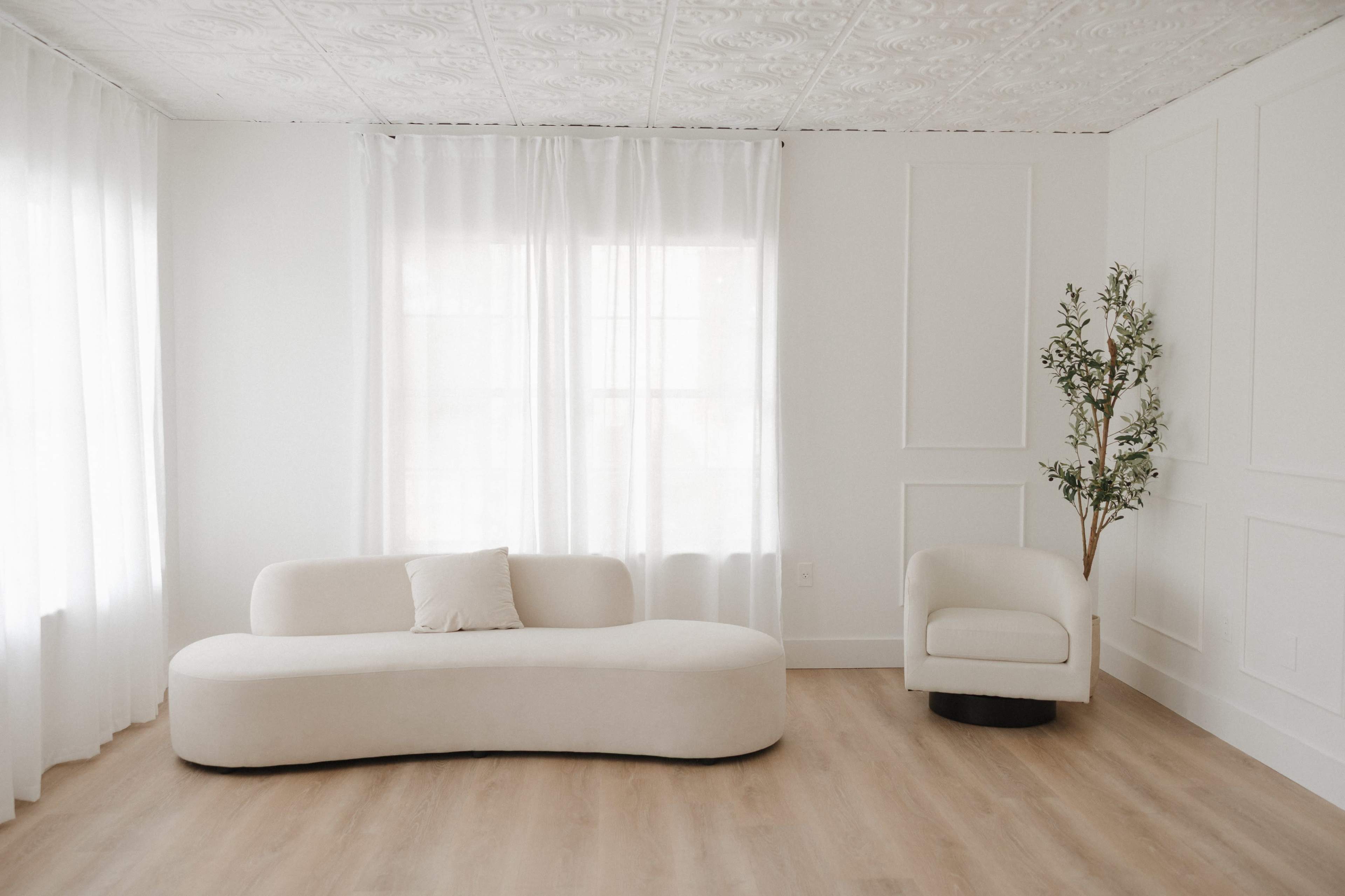 The image shows a minimalist living room featuring a curved beige sofa, a white armchair, large windows with sheer curtains, and a decorative potted plant.