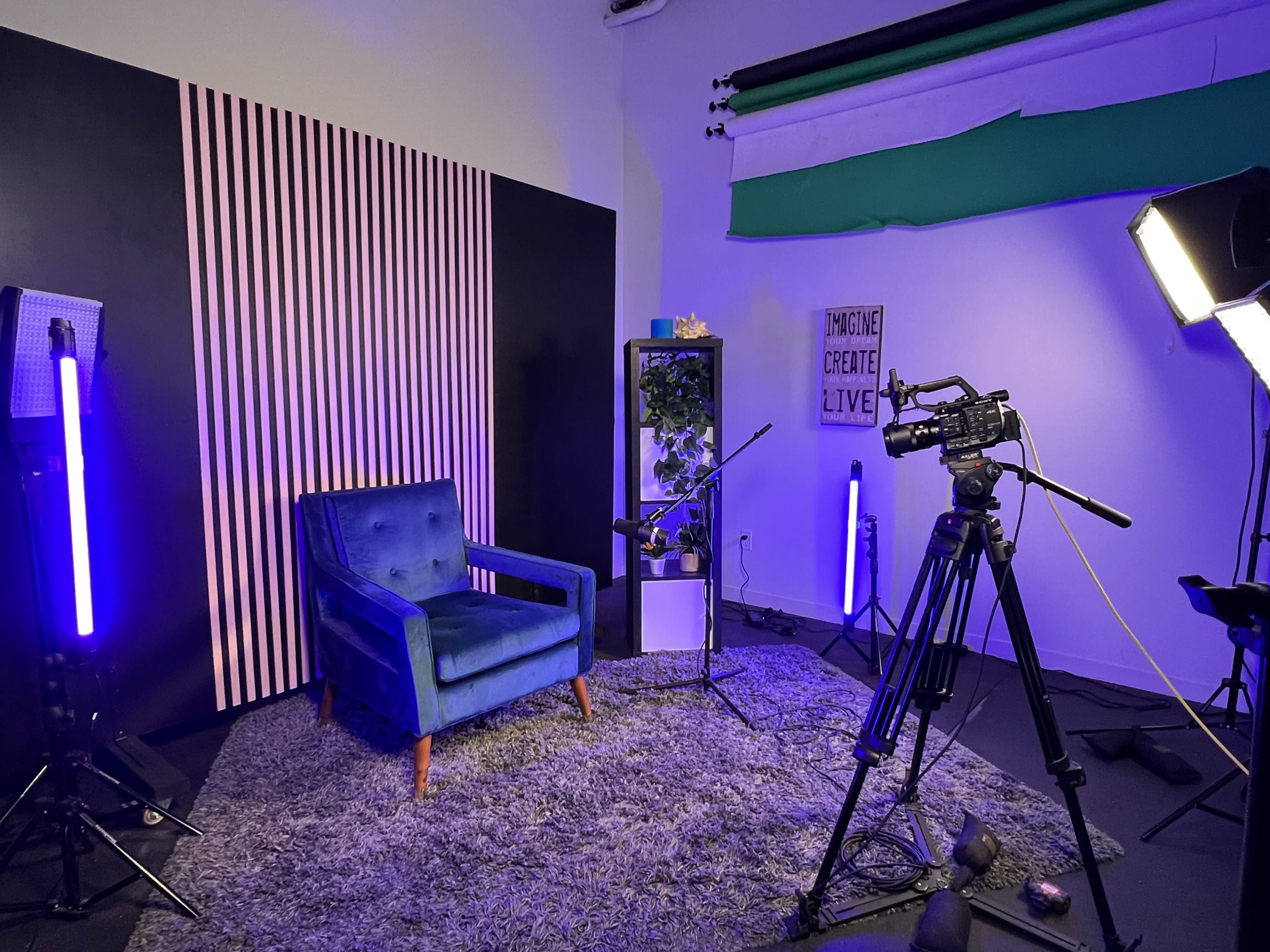 The scene features a blue chair on a gray rug in a studio setting, equipped with cameras and lighting, with a striped wall and a potted plant in the background.