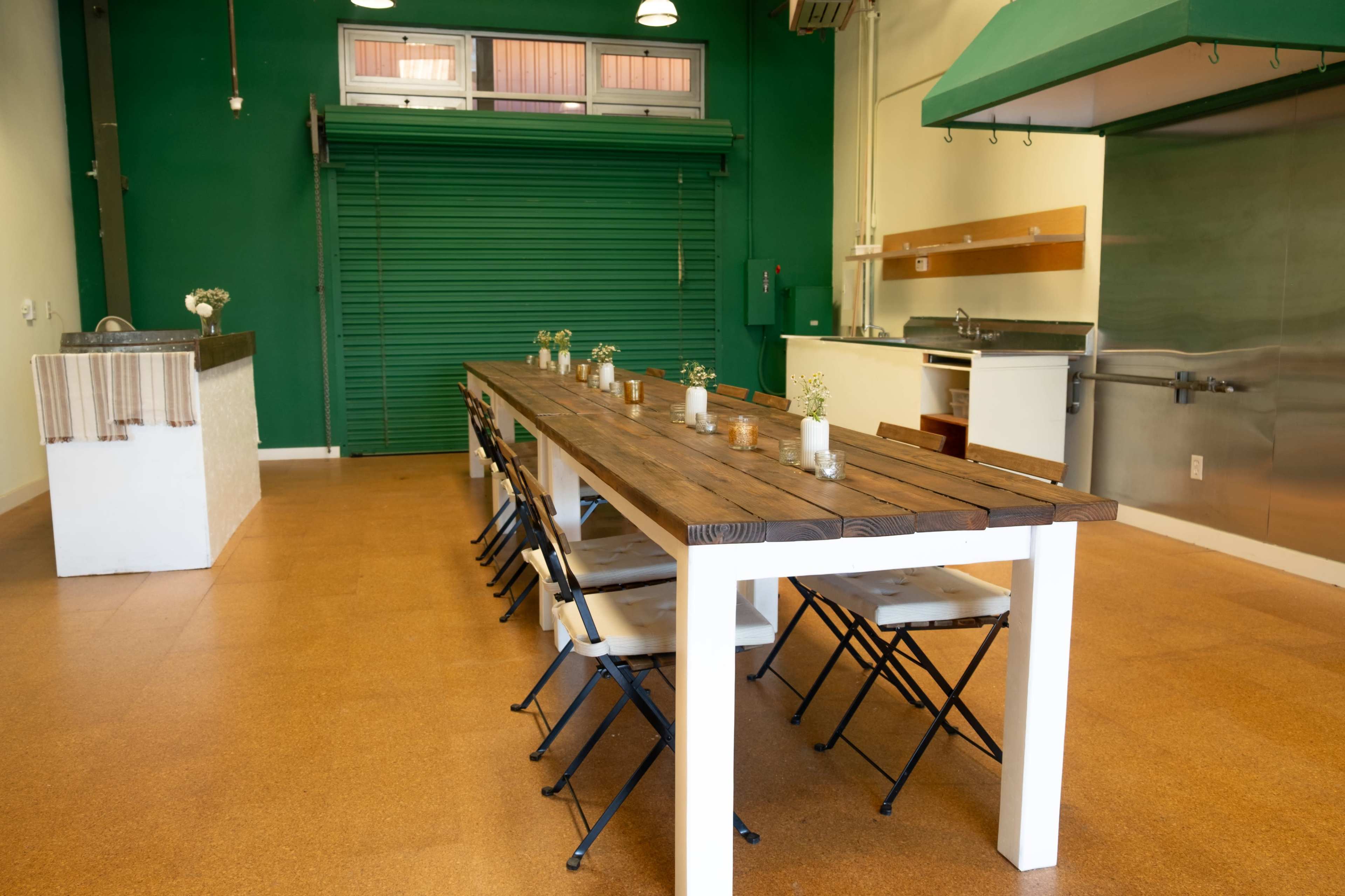 The image shows a spacious room with a long wooden table set with small vases, surrounded by folding chairs, and features green walls and a kitchen area with stainless steel appliances.