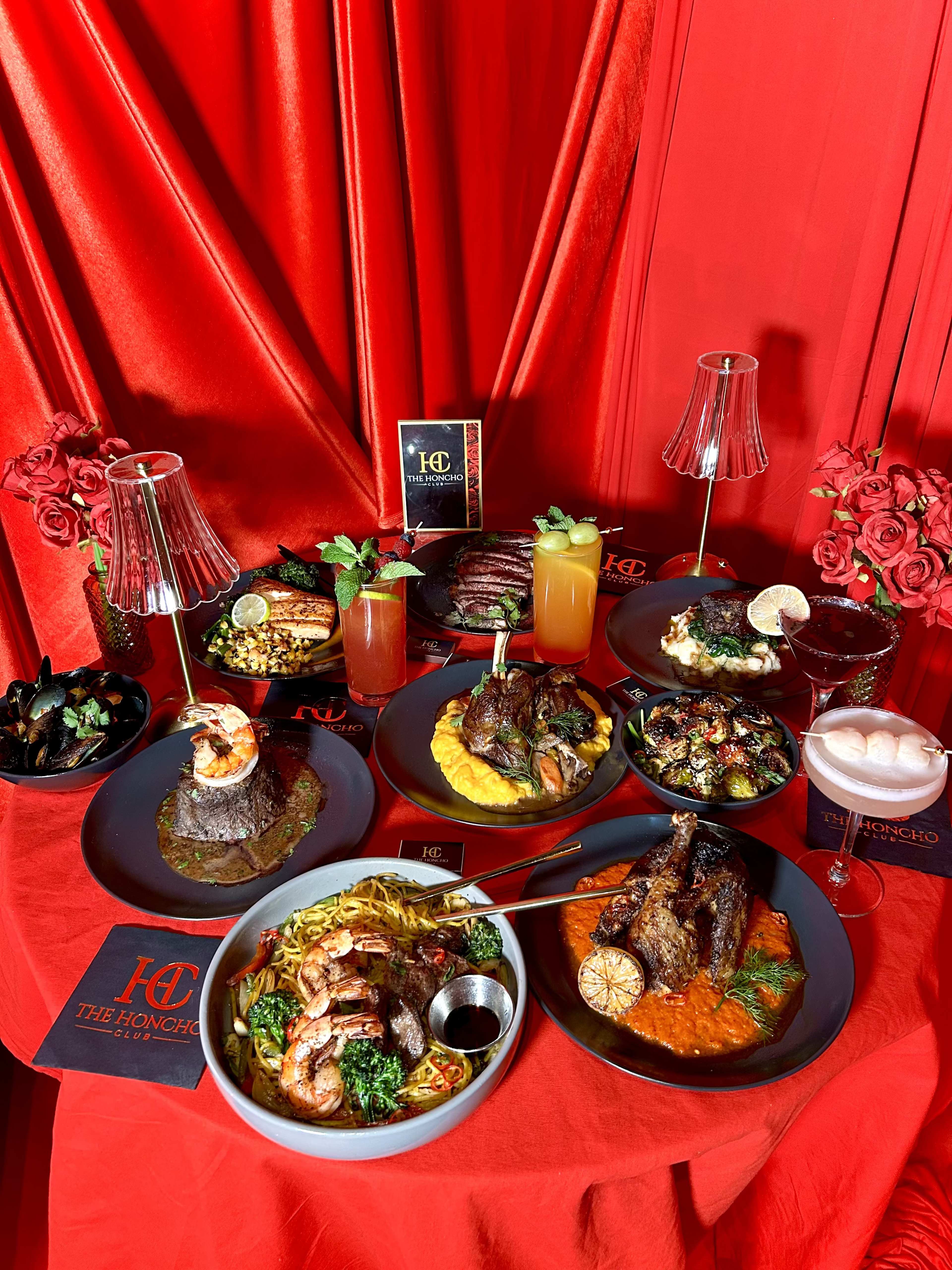 A table is covered with various gourmet dishes, including grilled meats, seafood, pasta, and vibrant sauces, all set against a red backdrop with decorative elements.