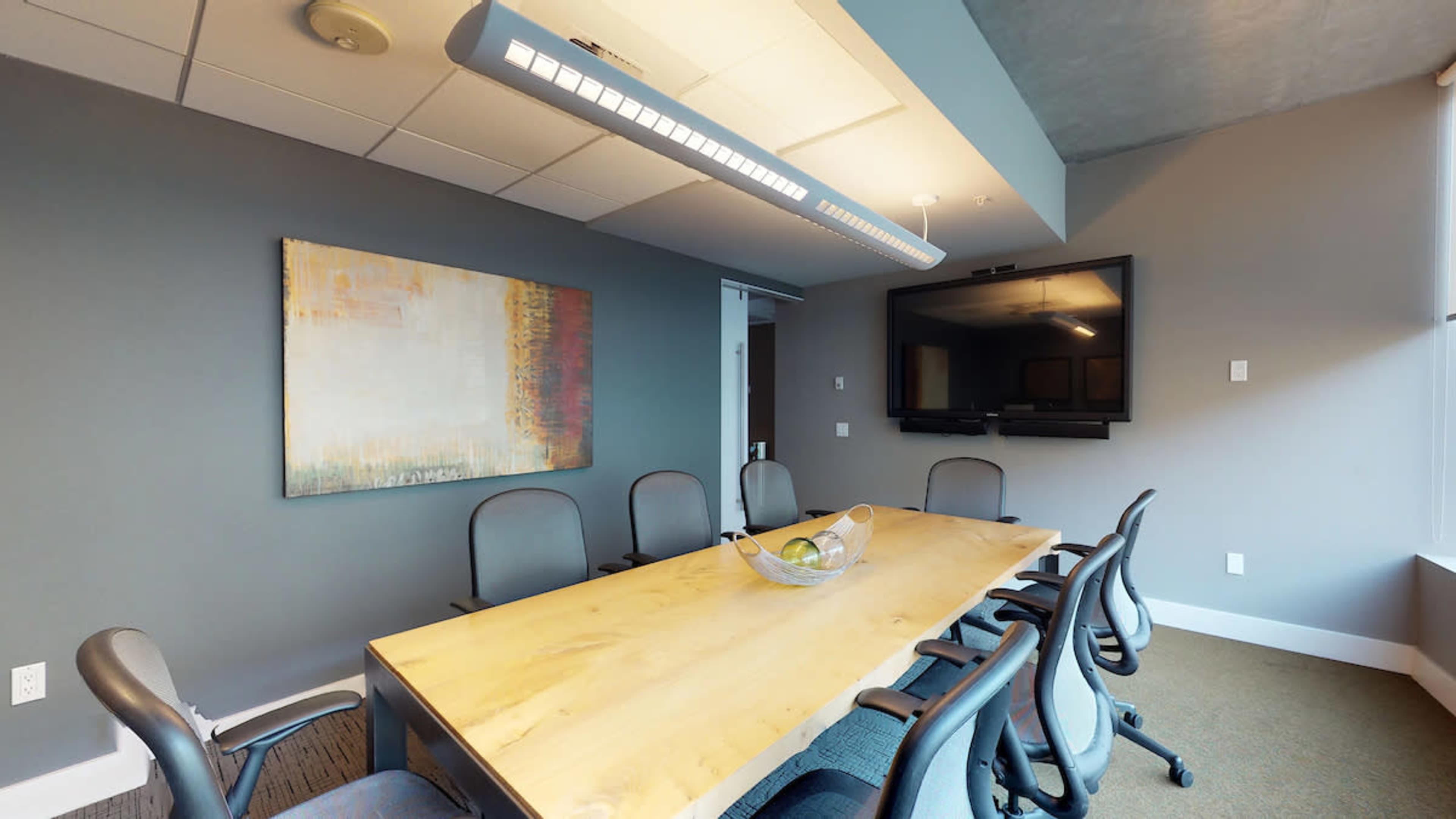 A meeting room features a long wooden table surrounded by ergonomic chairs, with a large screen mounted on the wall and a painting hanging nearby.