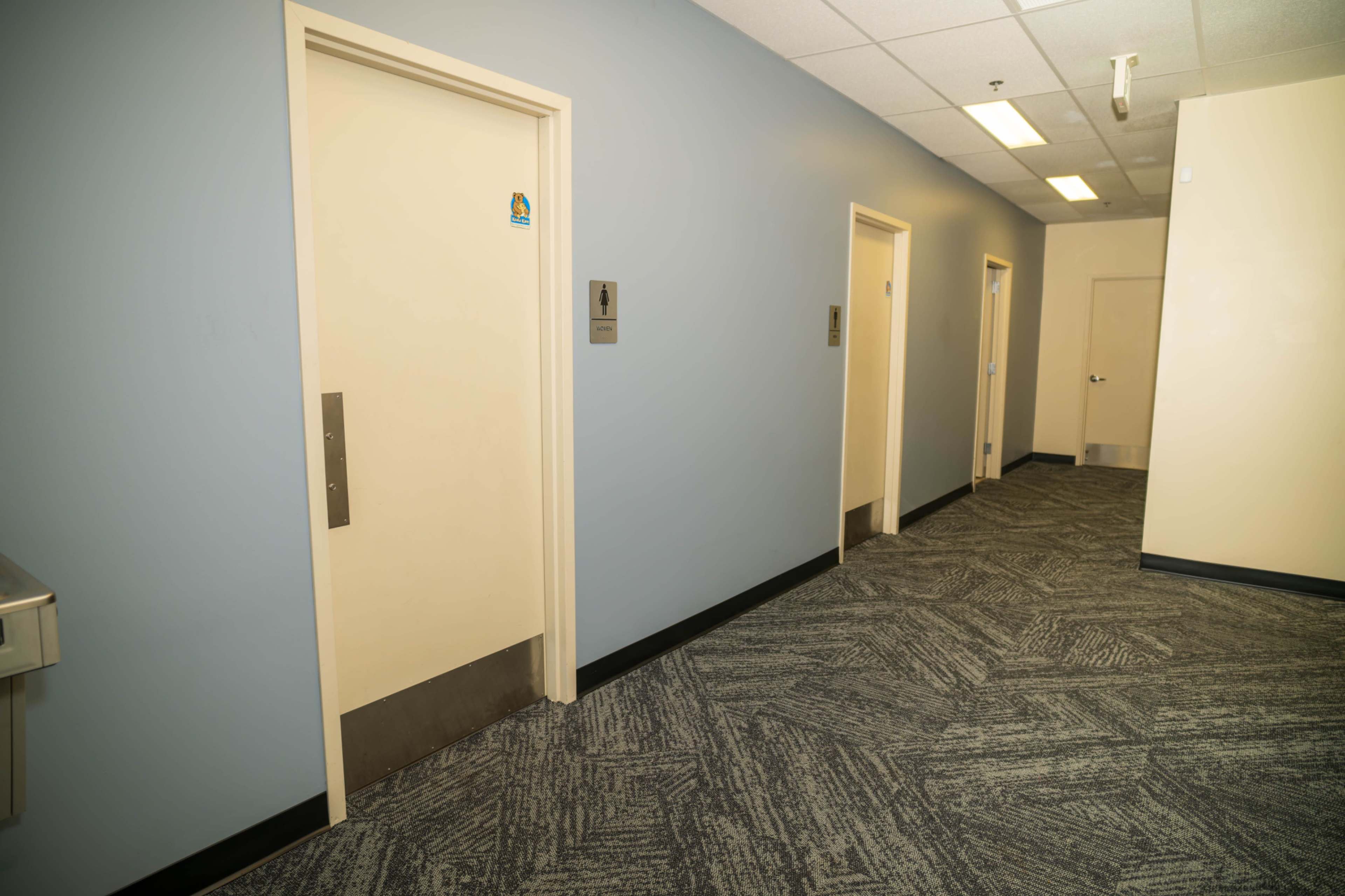 The image depicts a hallway with several closed doorways on one side and a drinking fountain on the opposite wall.