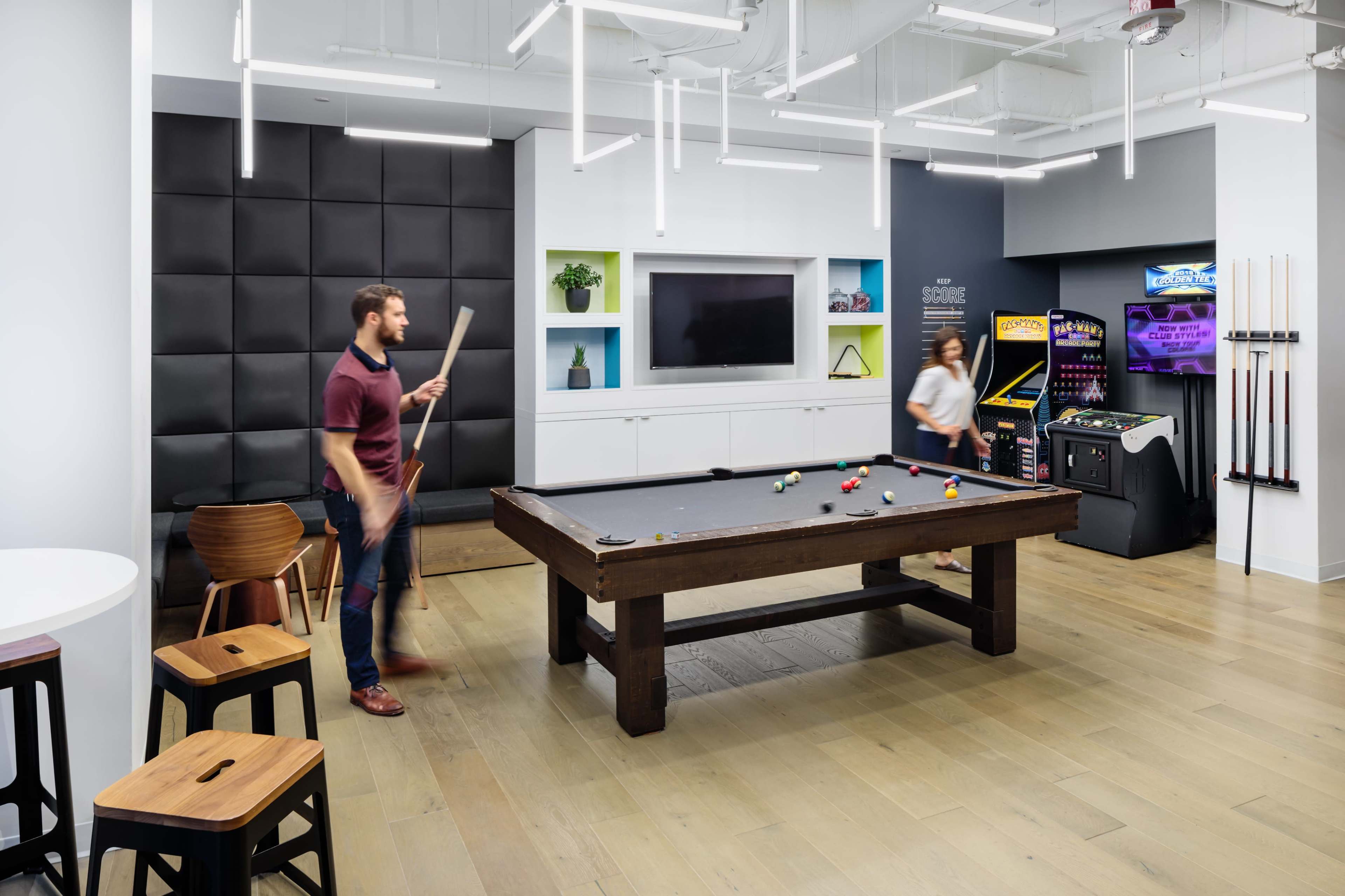 The scene features a modern game room with a billiards table, arcade machines, and a television, where one person is holding a cue stick while another is engaged with the arcade games.