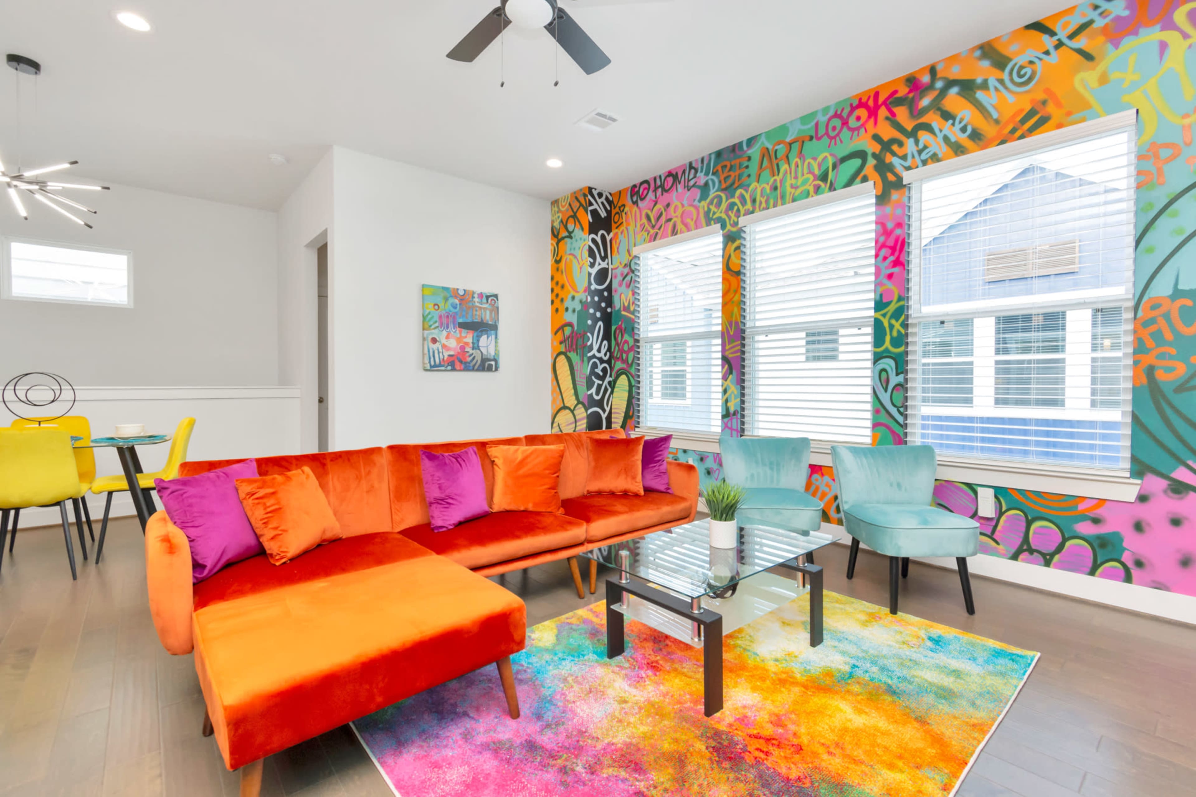 A modern living room features a bright orange sectional sofa, colorful accent pillows, a glass coffee table, and a vibrant graffiti wall.
