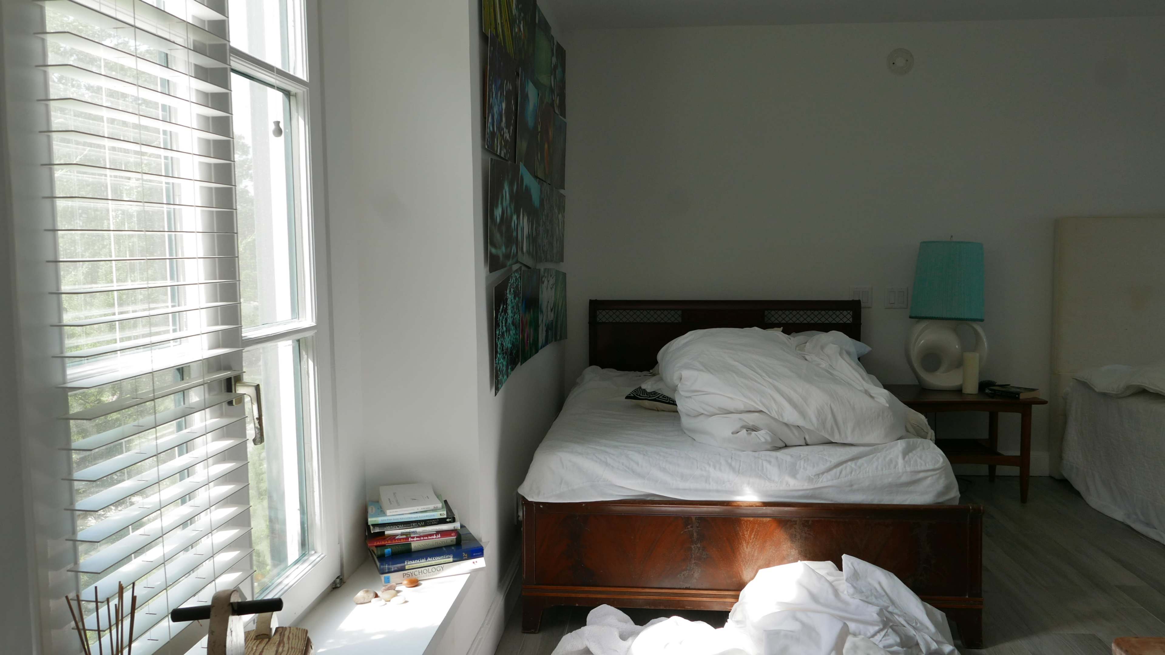 A bed with rumpled white sheets and a large painting on the wall is situated in a bright room next to a window with partially drawn blinds.