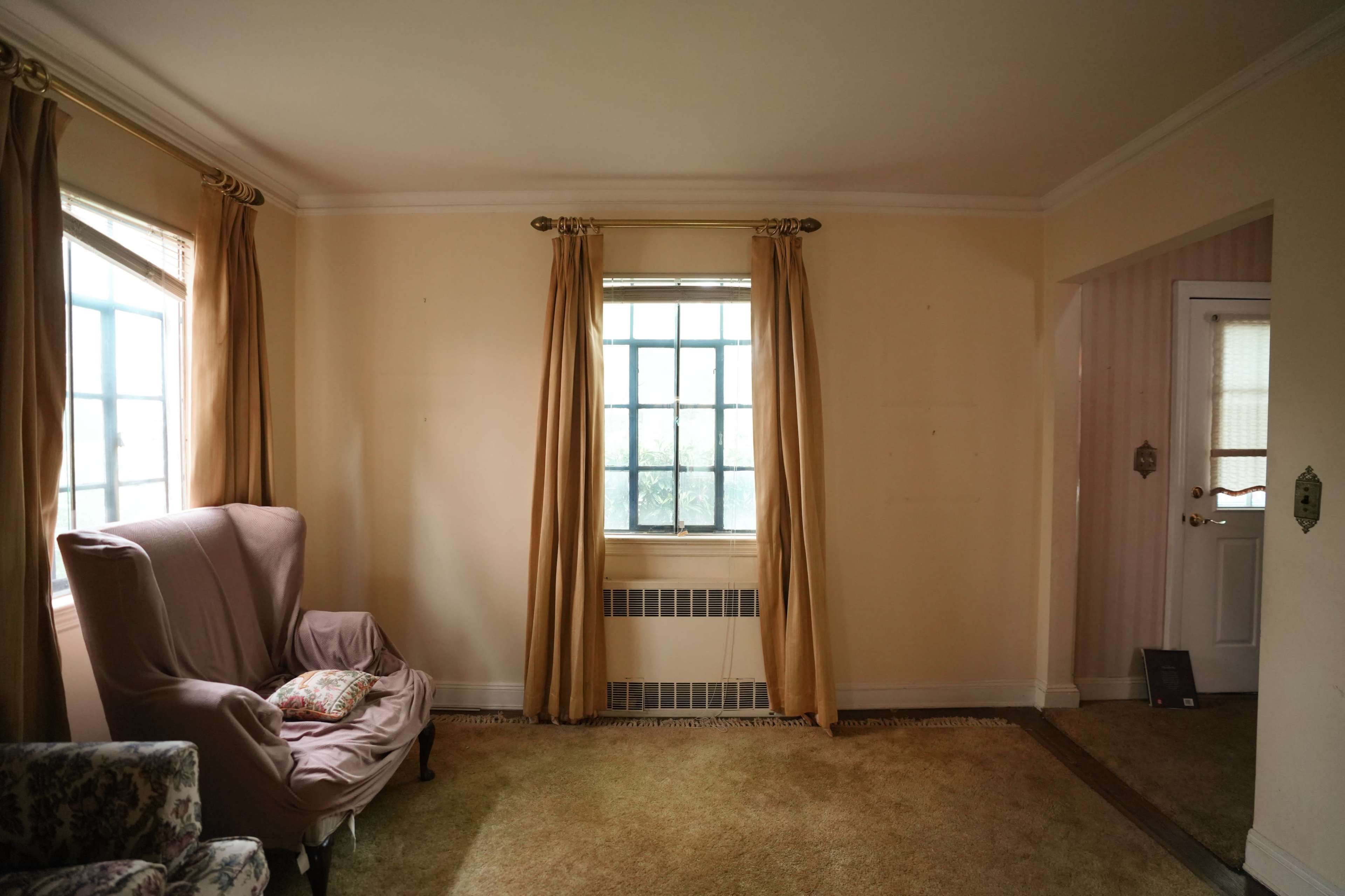 The room features a pink armchair, beige walls, large windows with drapes, and a door leading to another space.