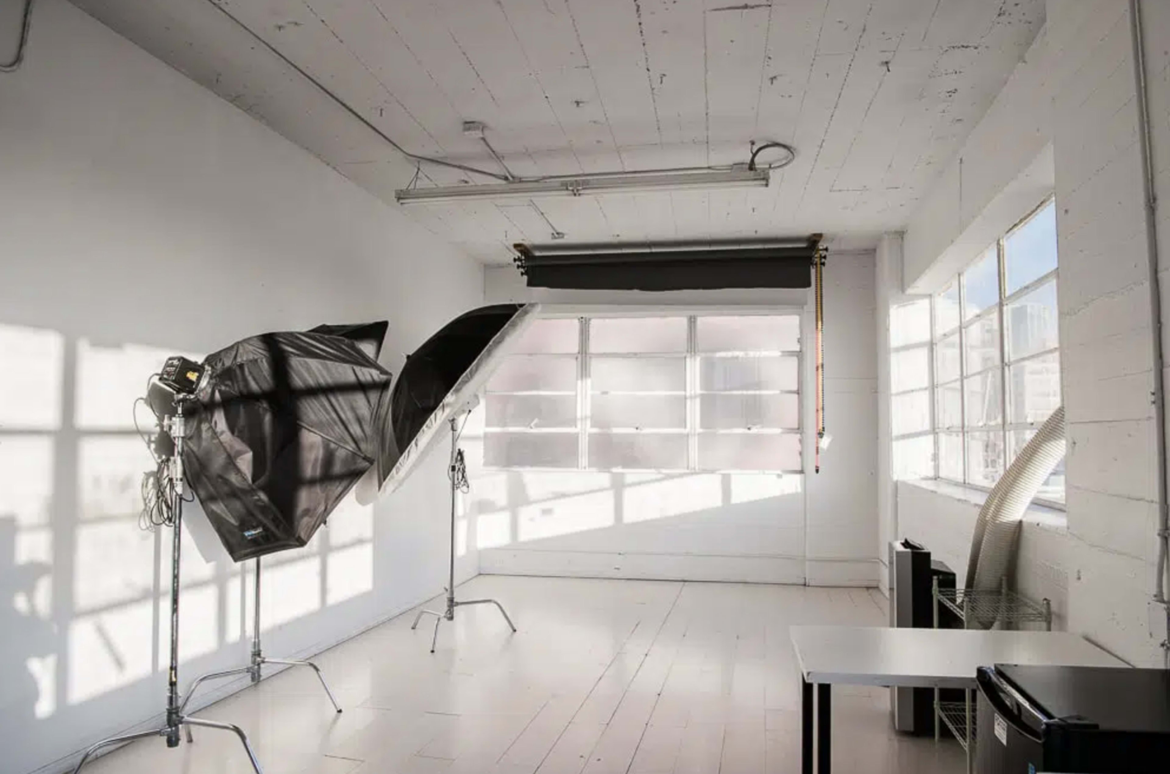 The image shows a well-lit photography studio with two softboxes positioned near a large window and a table in the corner.