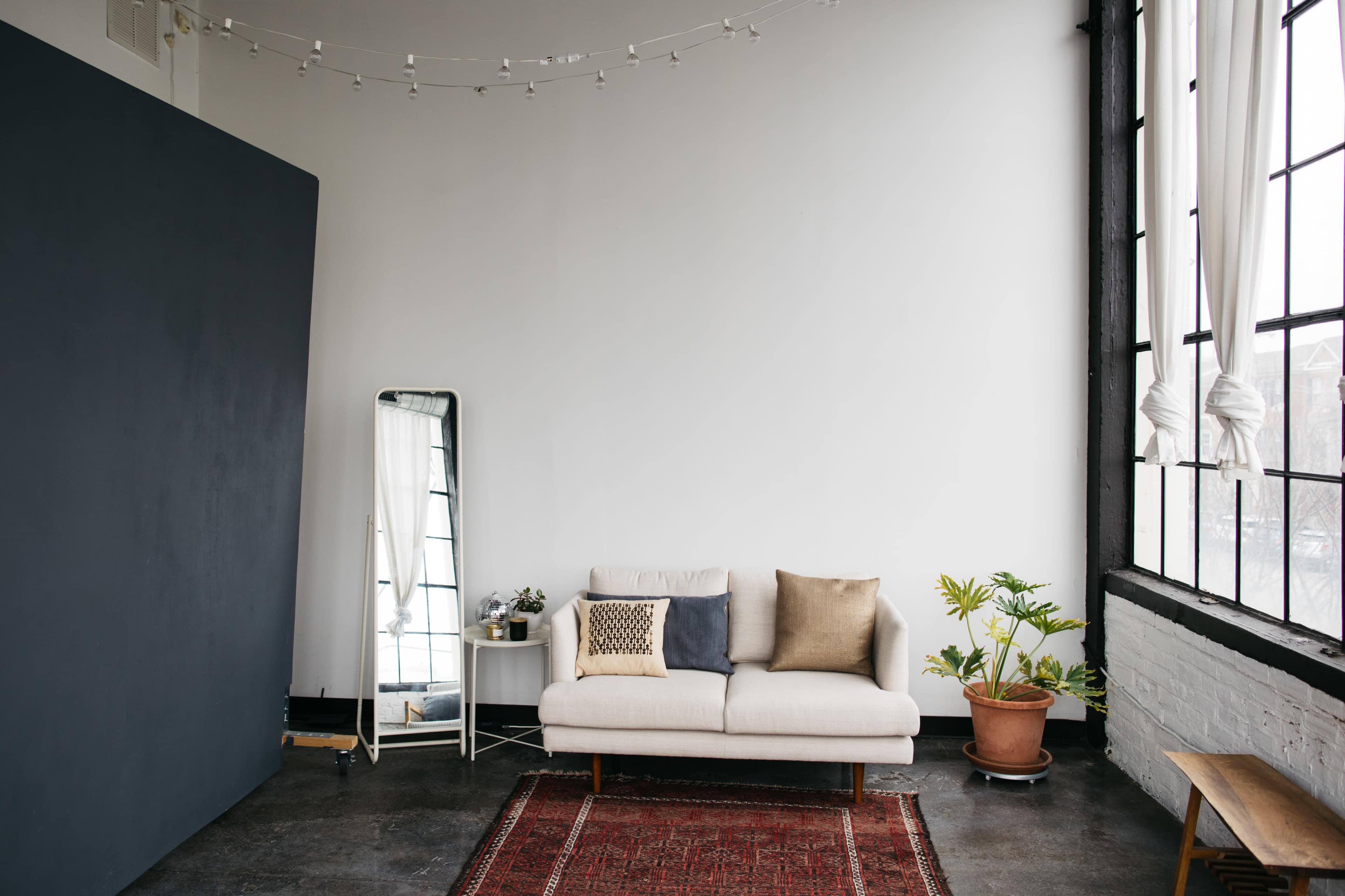 The image shows a minimalist living space with a light-colored sofa, a large mirror, a potted plant, and a patterned rug on a concrete floor.