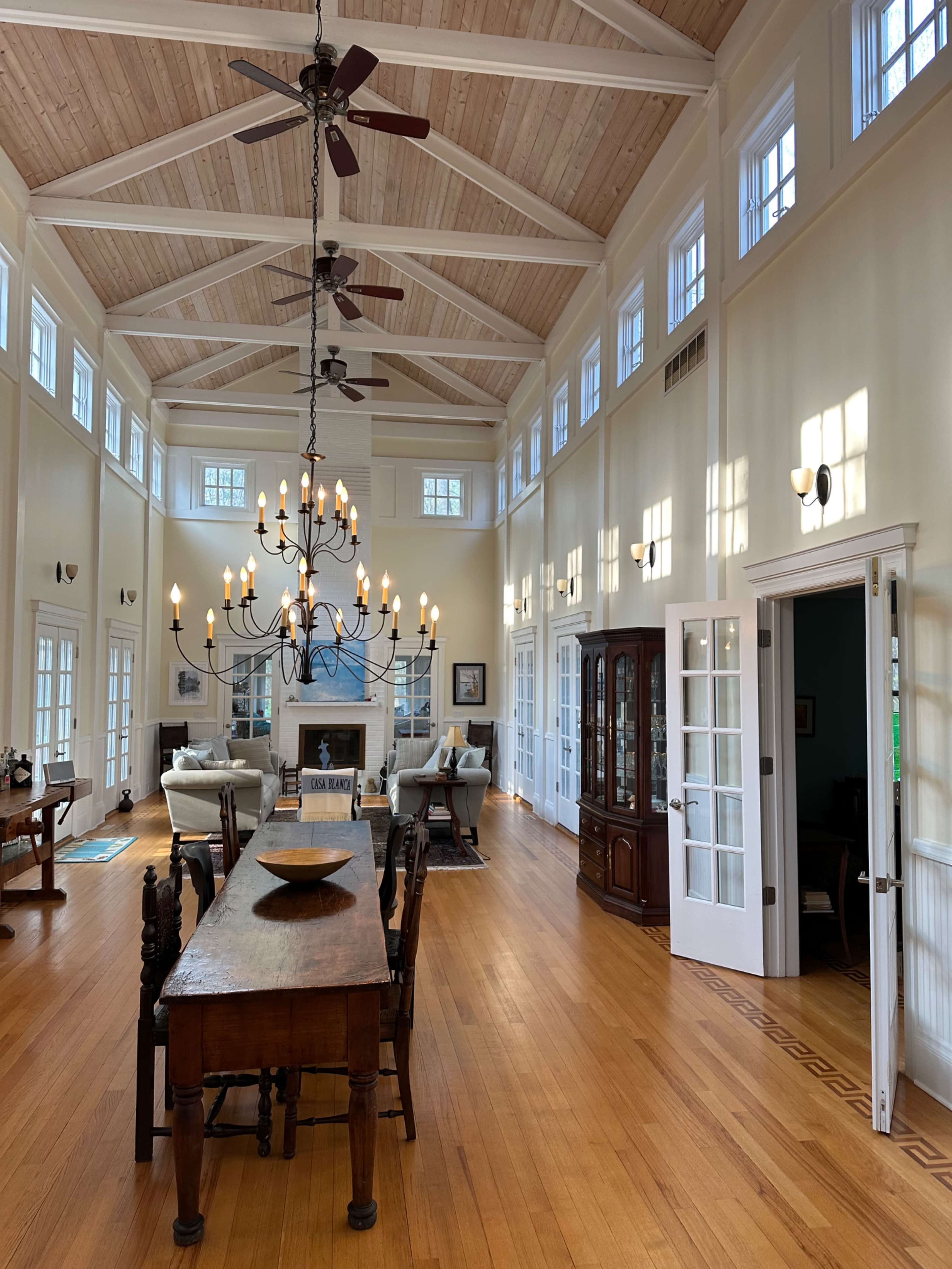 A spacious, well-lit interior with high ceilings, wooden beams, a long dining table, and a chandelier, leading to an adjoining room.