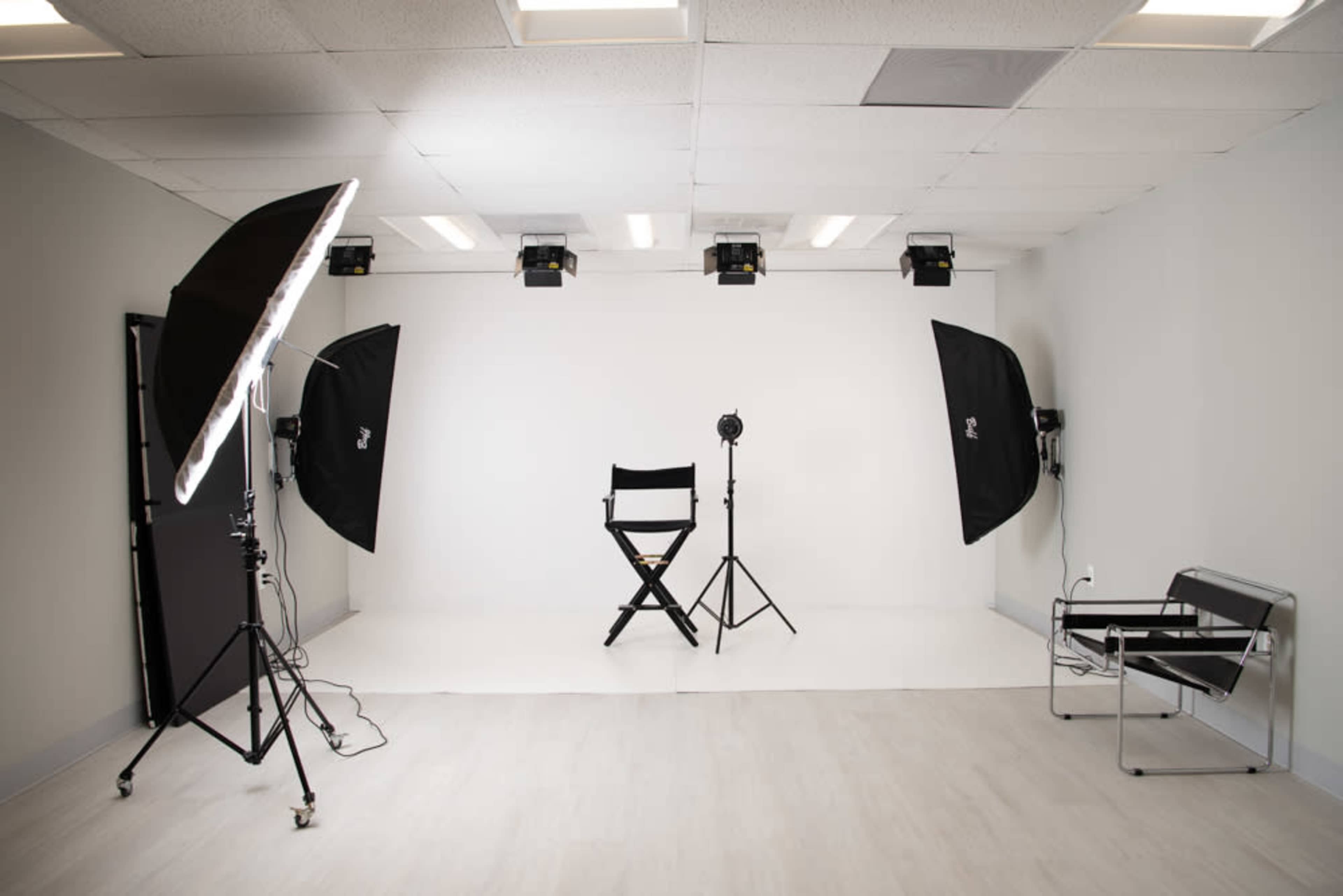The image depicts a photography studio with a white backdrop, a director's chair, and various lighting equipment arranged around the space.