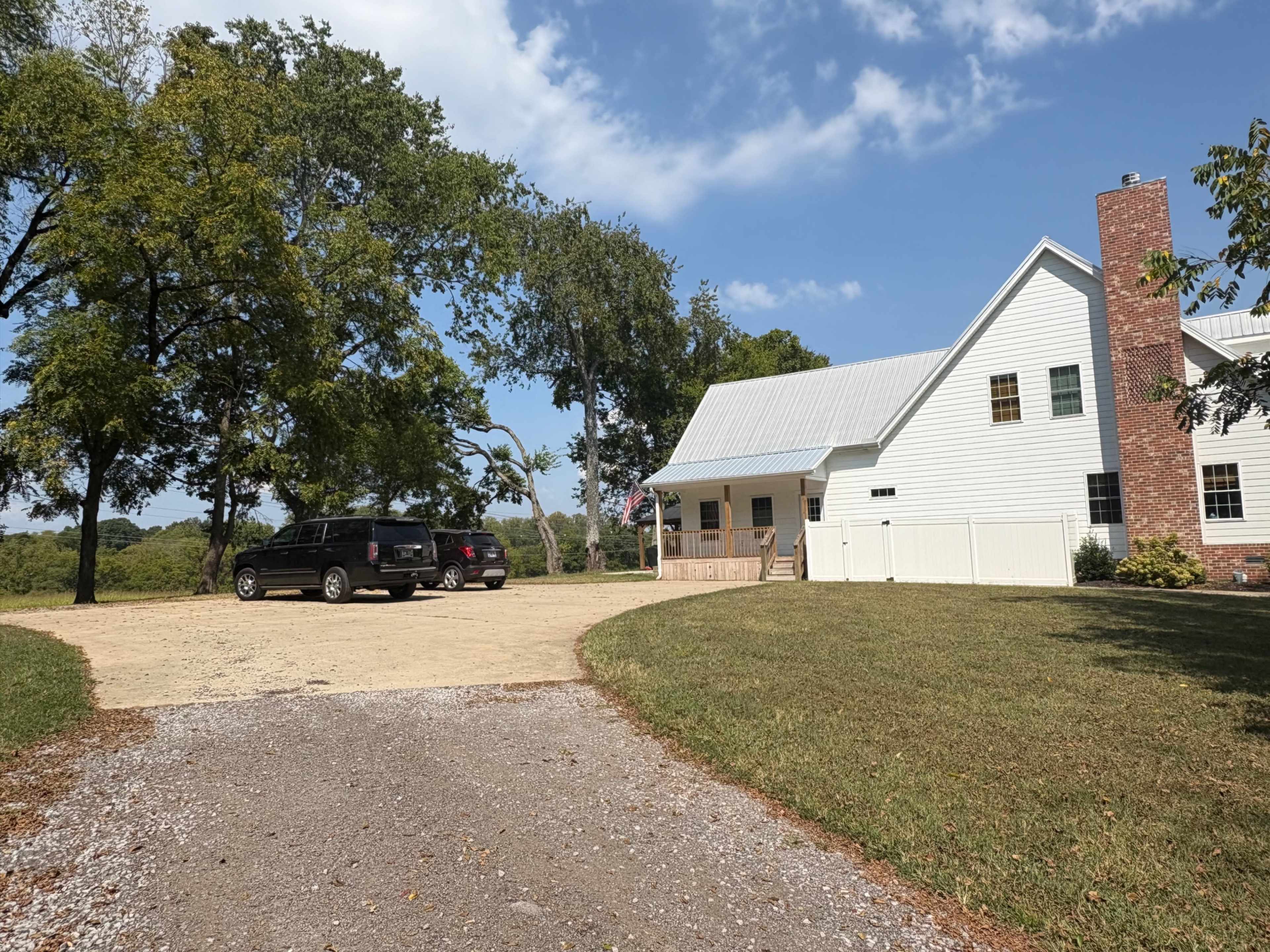 The image shows a driveway leading to a white house with a metal roof, surrounded by trees and featuring parked vehicles on the gravel and dirt surface.