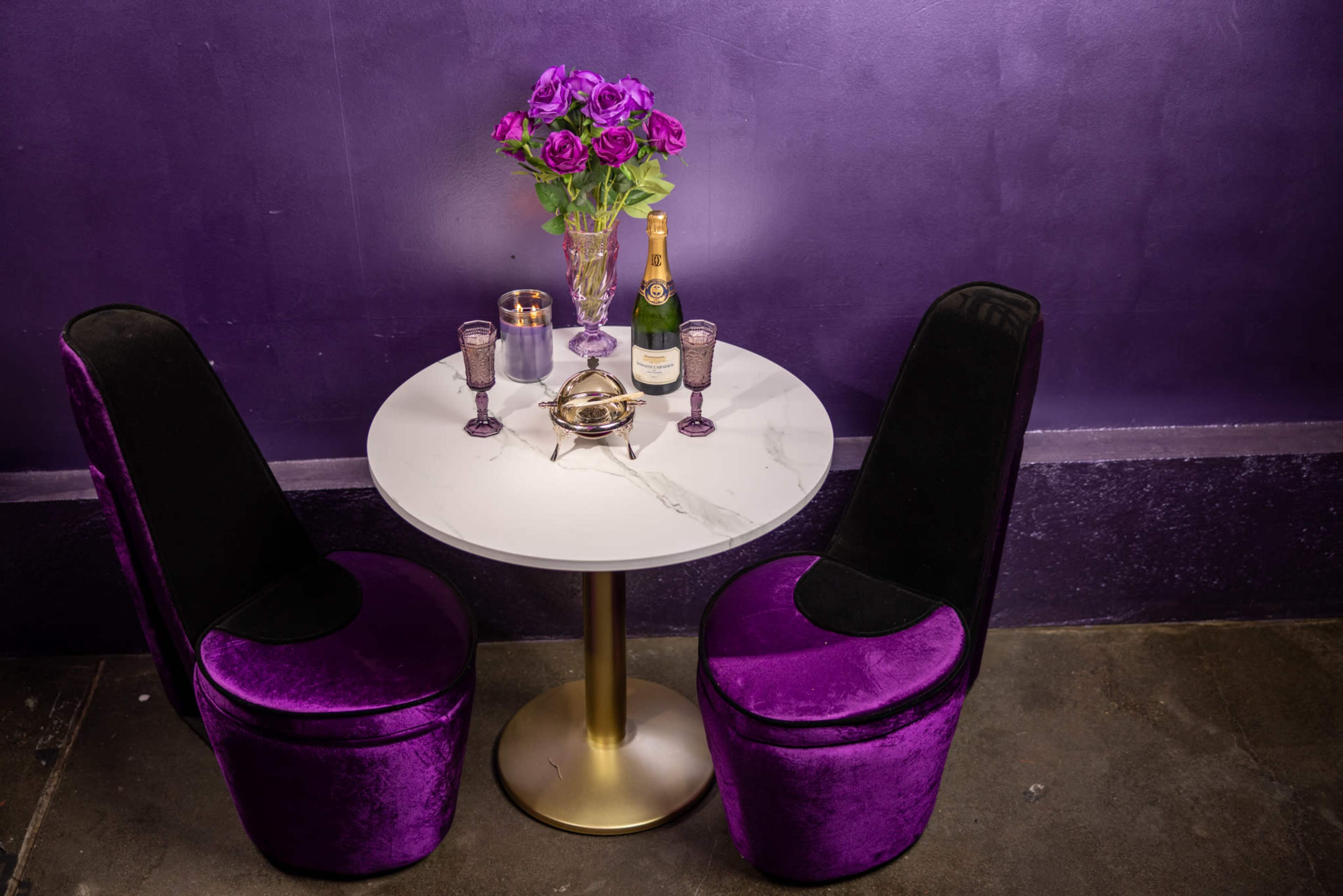 A circular marble table with a gold base is set between two purple upholstered chairs, featuring a vase of purple roses and a bottle of champagne.