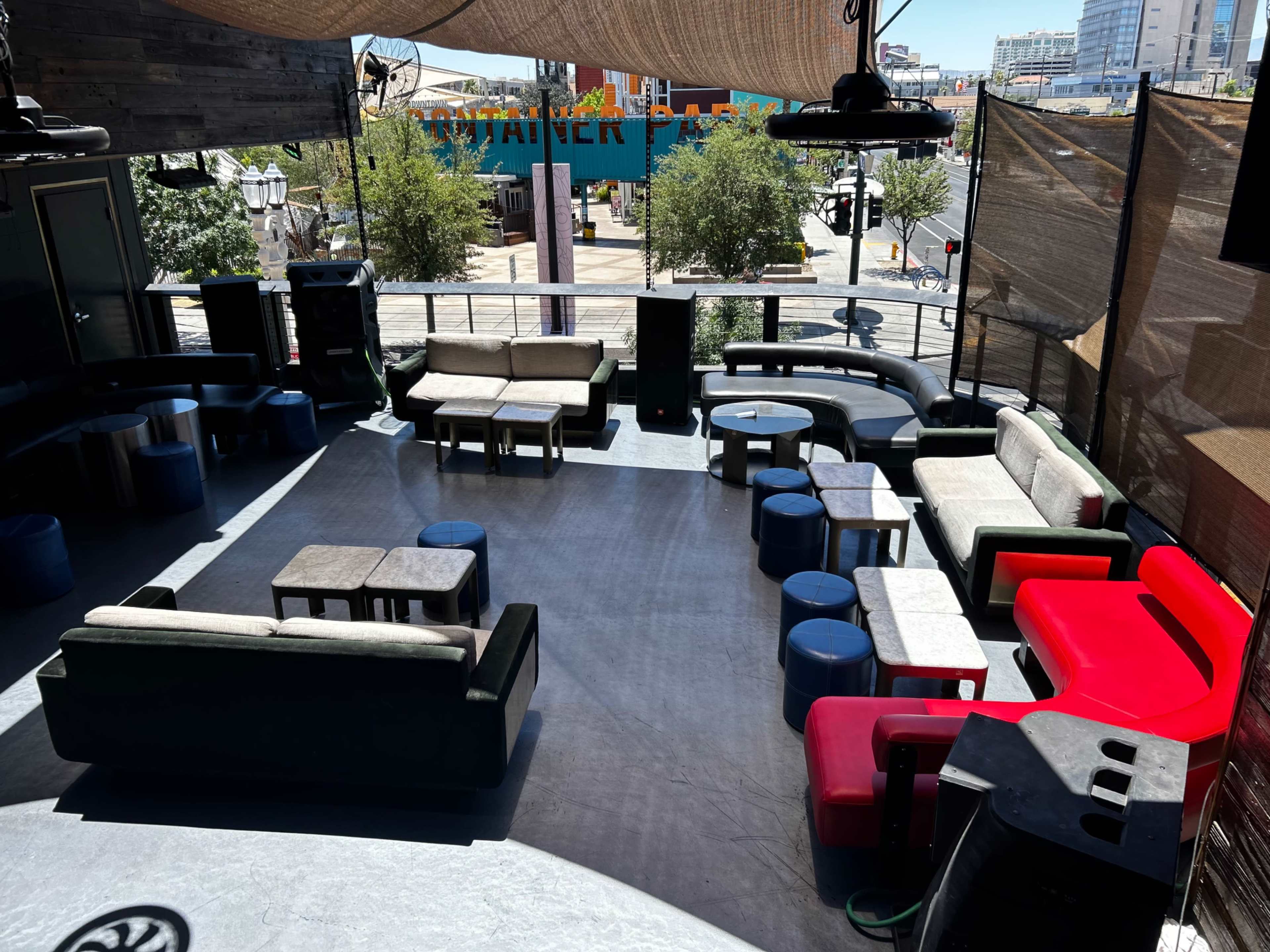 The image shows an elevated outdoor lounge area featuring various seating arrangements, including couches, stools, and tables, under a canopy.