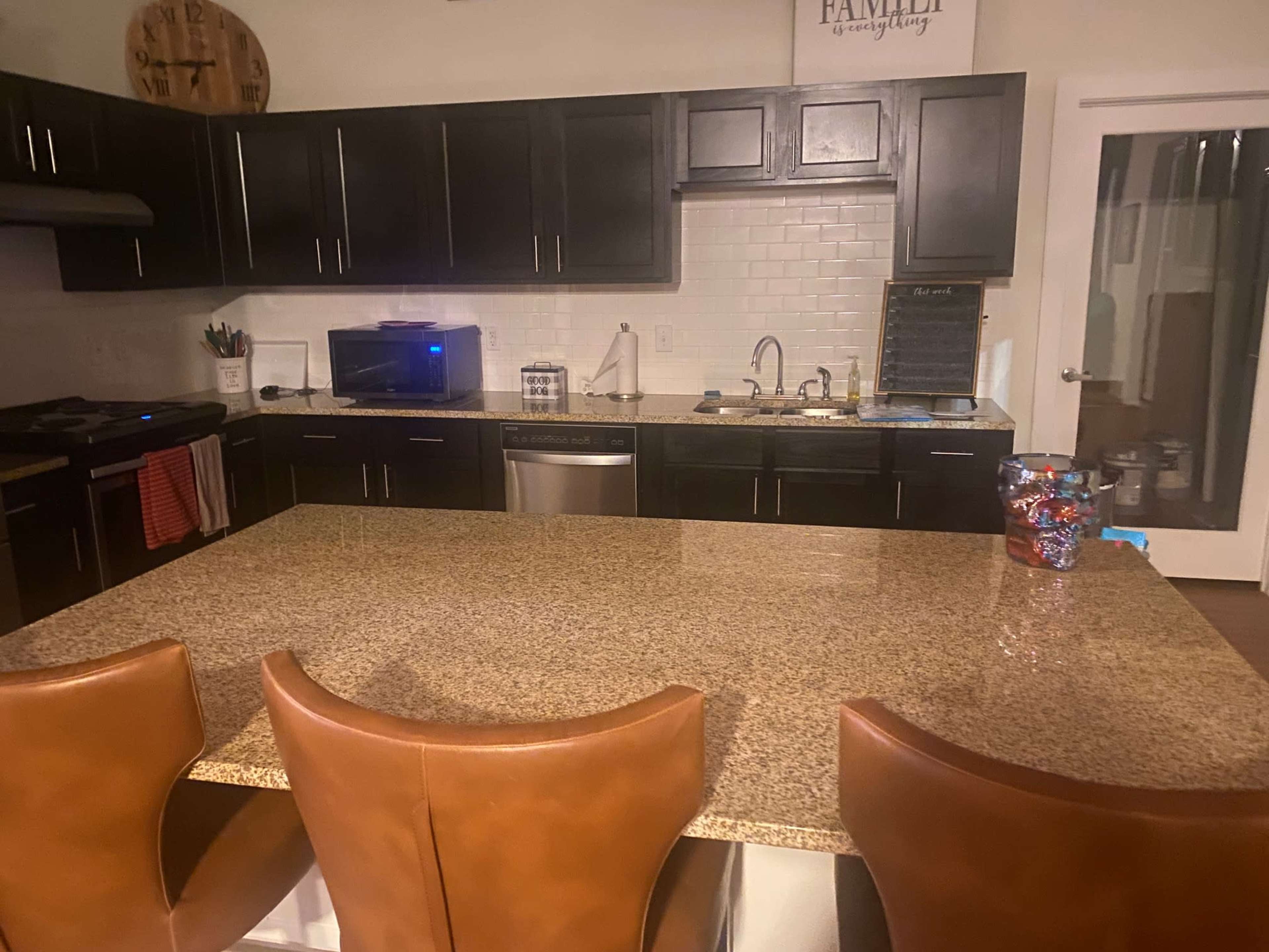 A modern kitchen features dark cabinets, a large granite island with four brown leather stools, and stainless steel appliances.