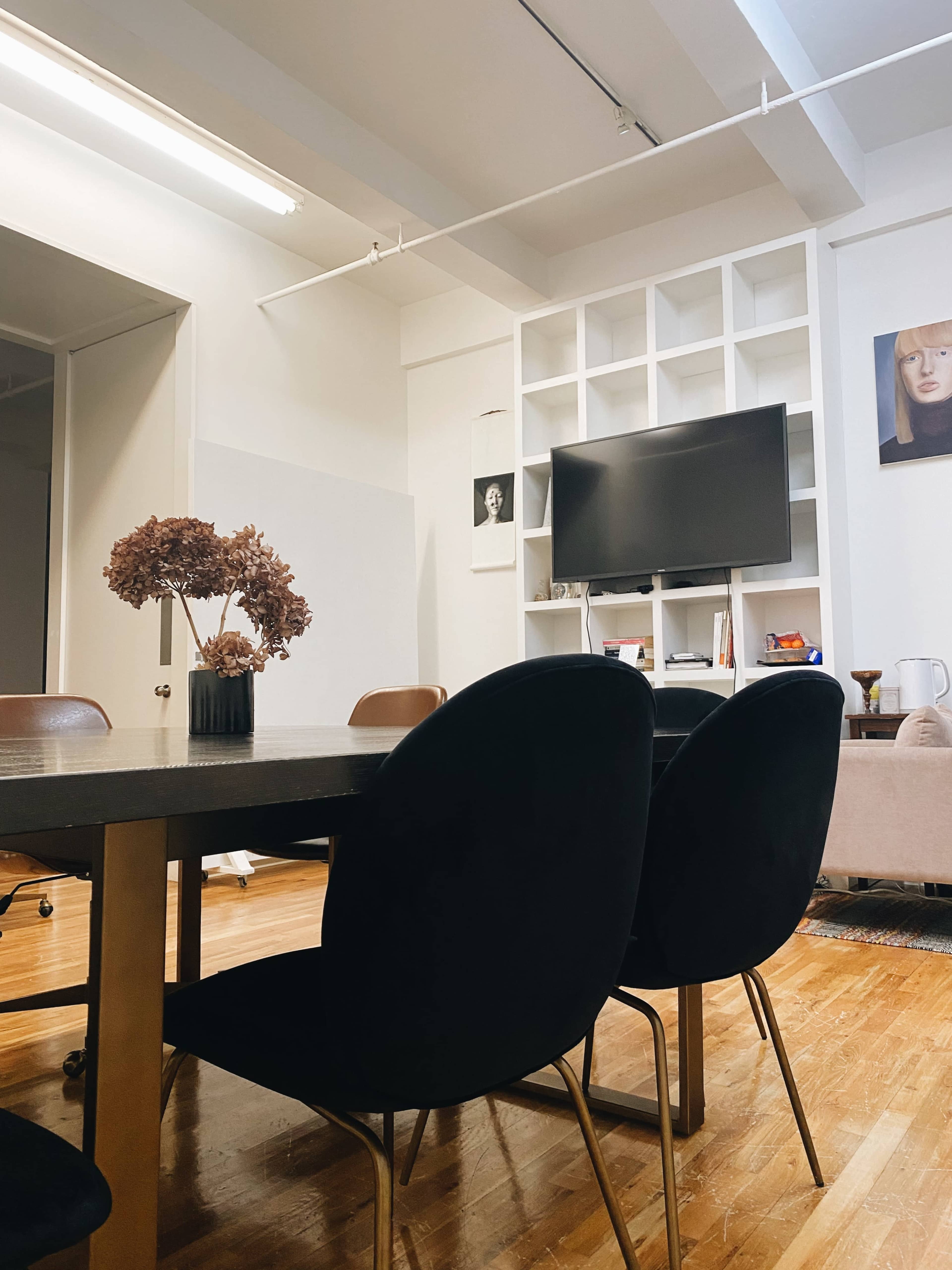 A modern meeting room features a wooden table surrounded by black chairs, with a TV mounted on the wall and decorative items on shelves.