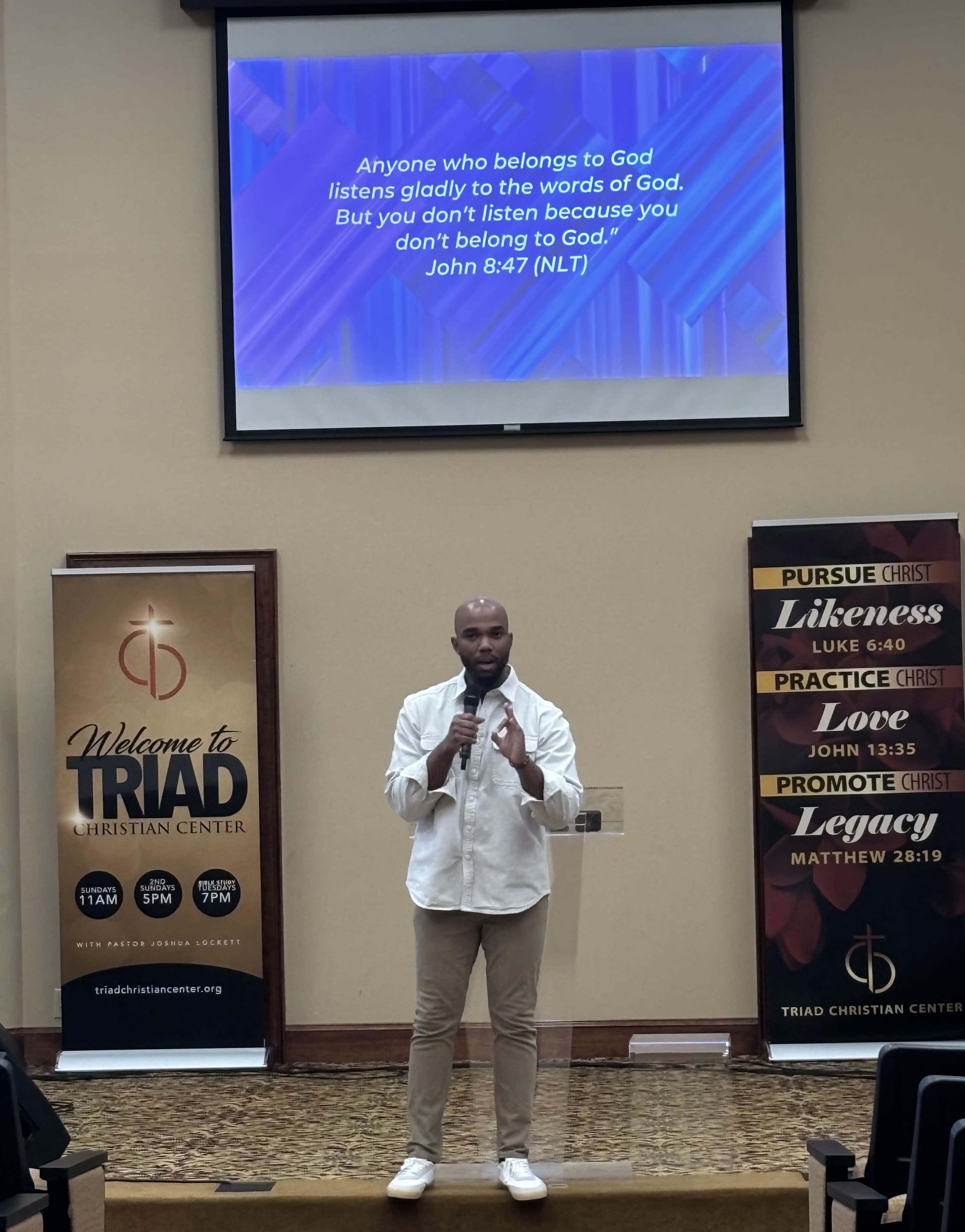A speaker stands in front of a screen displaying a Bible verse, with banners on either side promoting a Christian center.