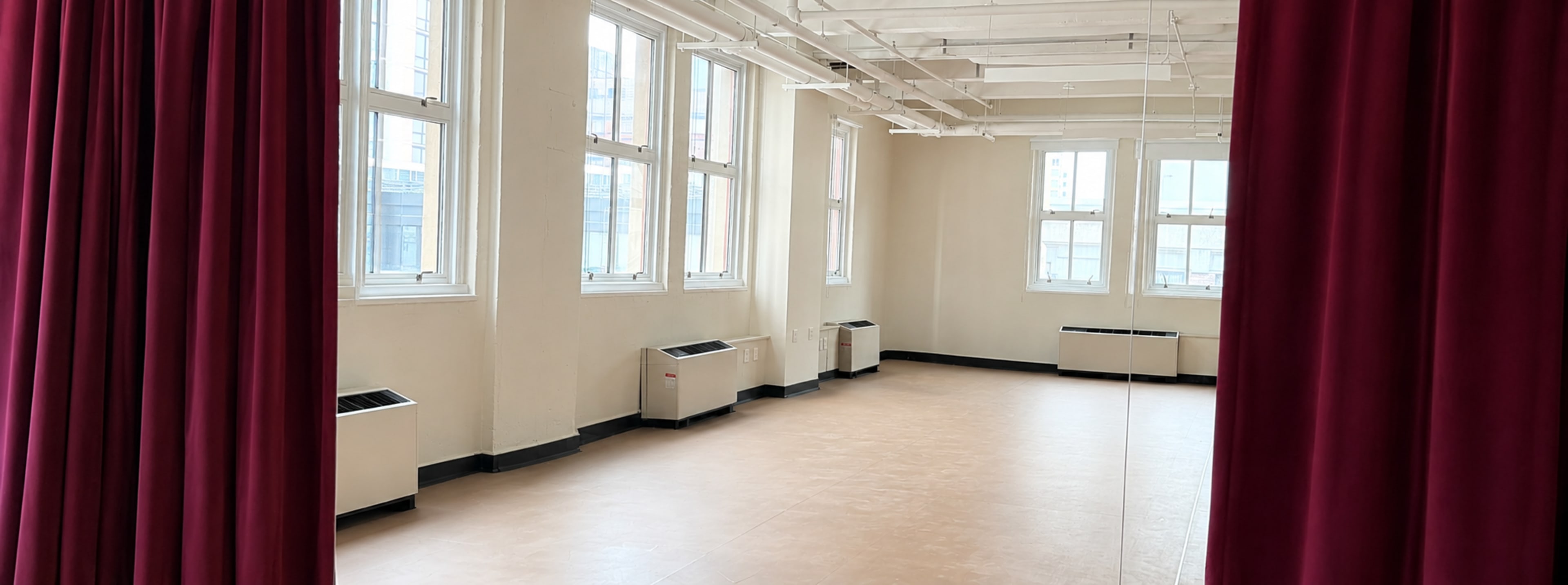 The image shows a bright, empty studio space with large windows, beige walls, and a mirrored wall, framed by red curtains.