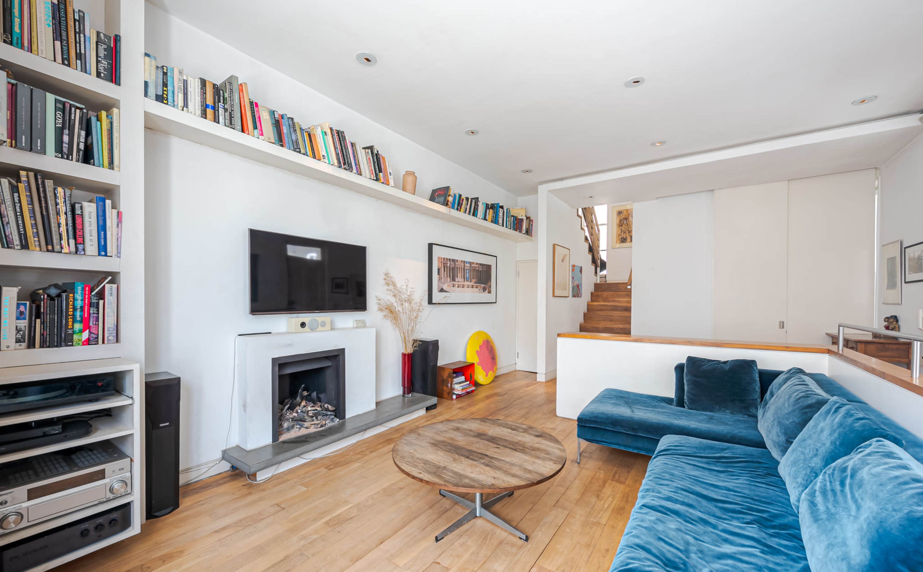 A living room features a blue sectional sofa, a wooden coffee table, a television mounted on the wall, and bookshelves filled with books along the upper wall.