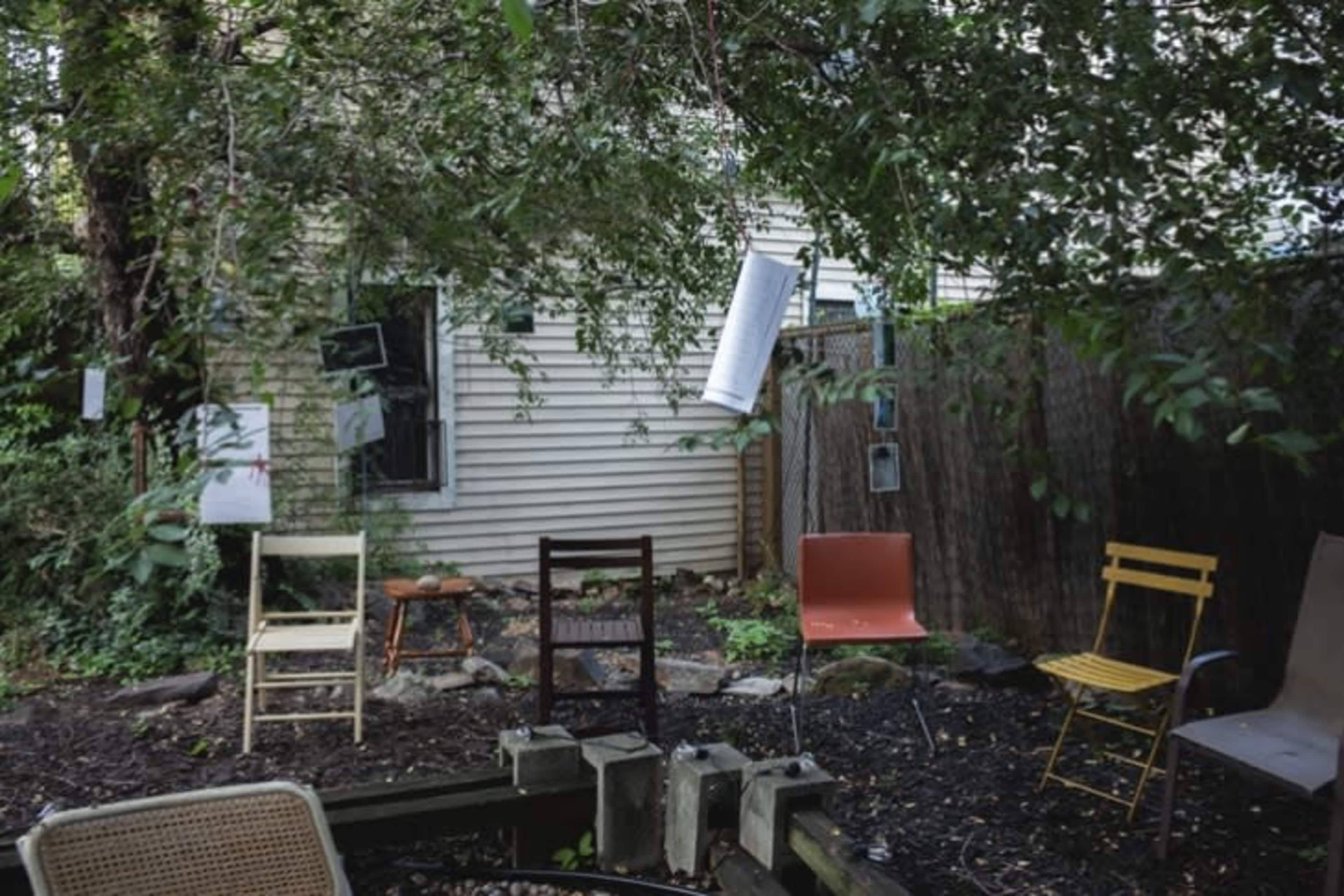A small backyard features several empty chairs arranged around a grassy area, with tree branches overhead and a house in the background.