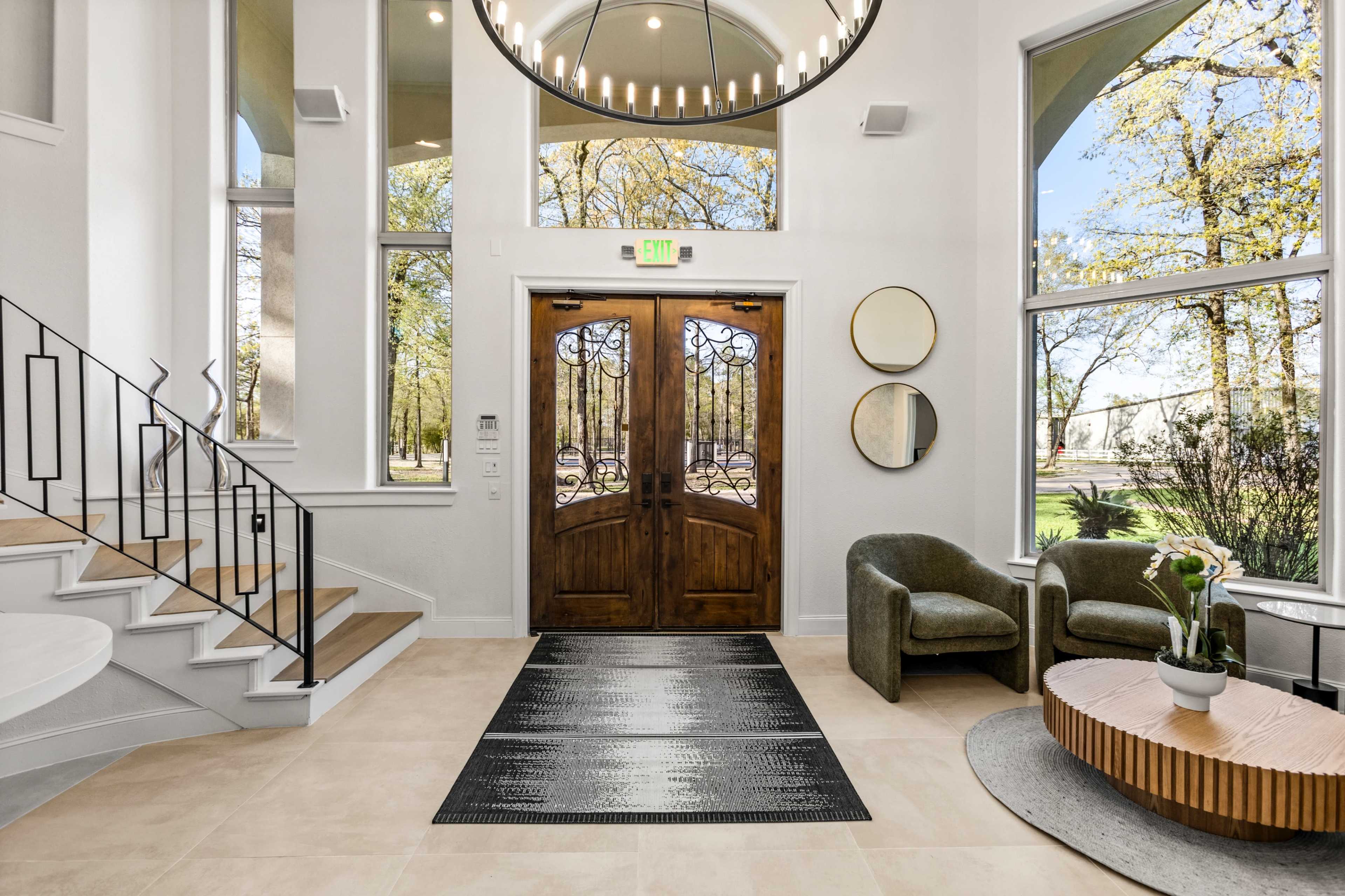 The image shows a spacious entryway with large double doors, a curved staircase, and modern seating arrangements near large windows.