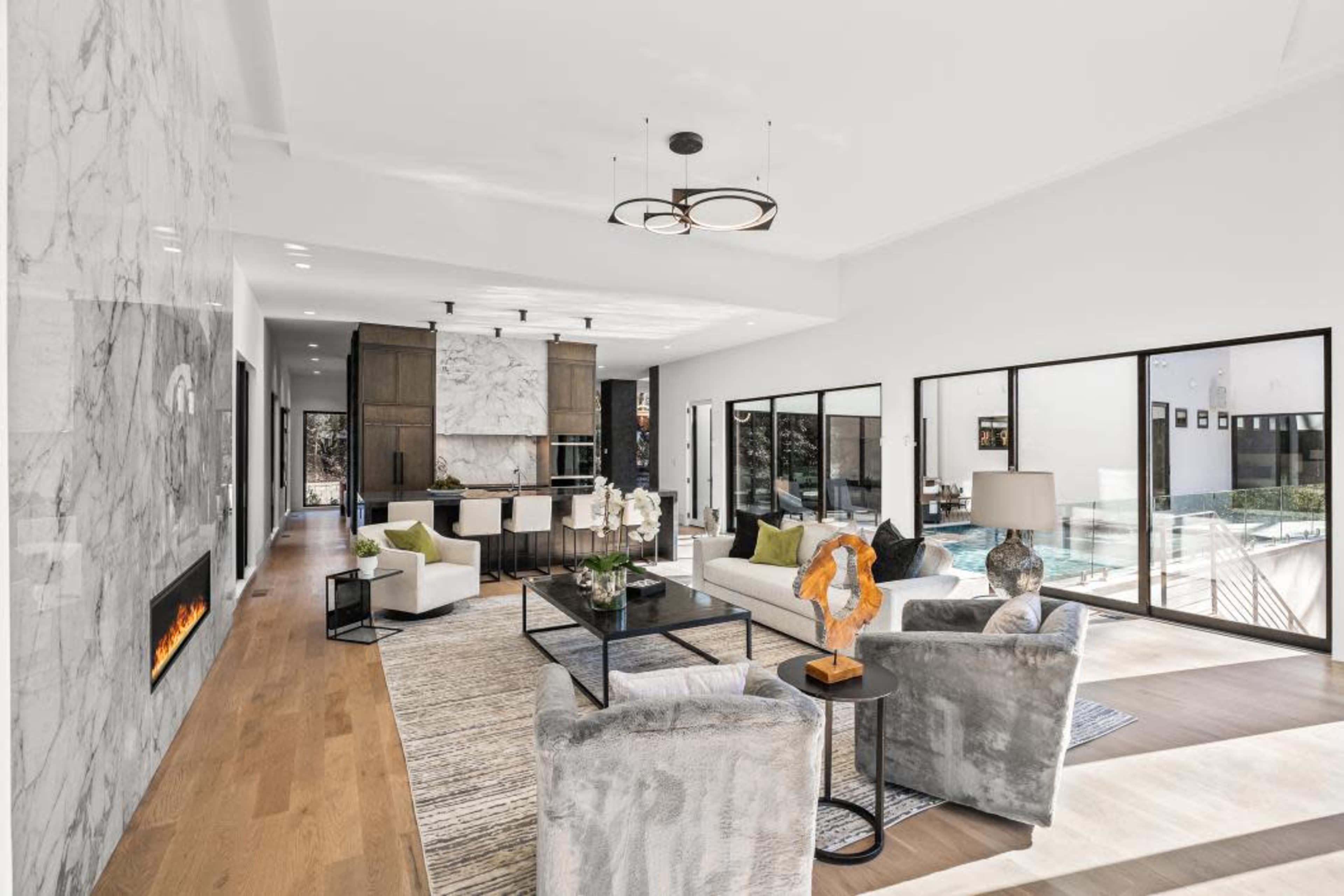 The image depicts a modern open-concept living area featuring a marble accent wall, a sleek fireplace, and large windows overlooking a pool.