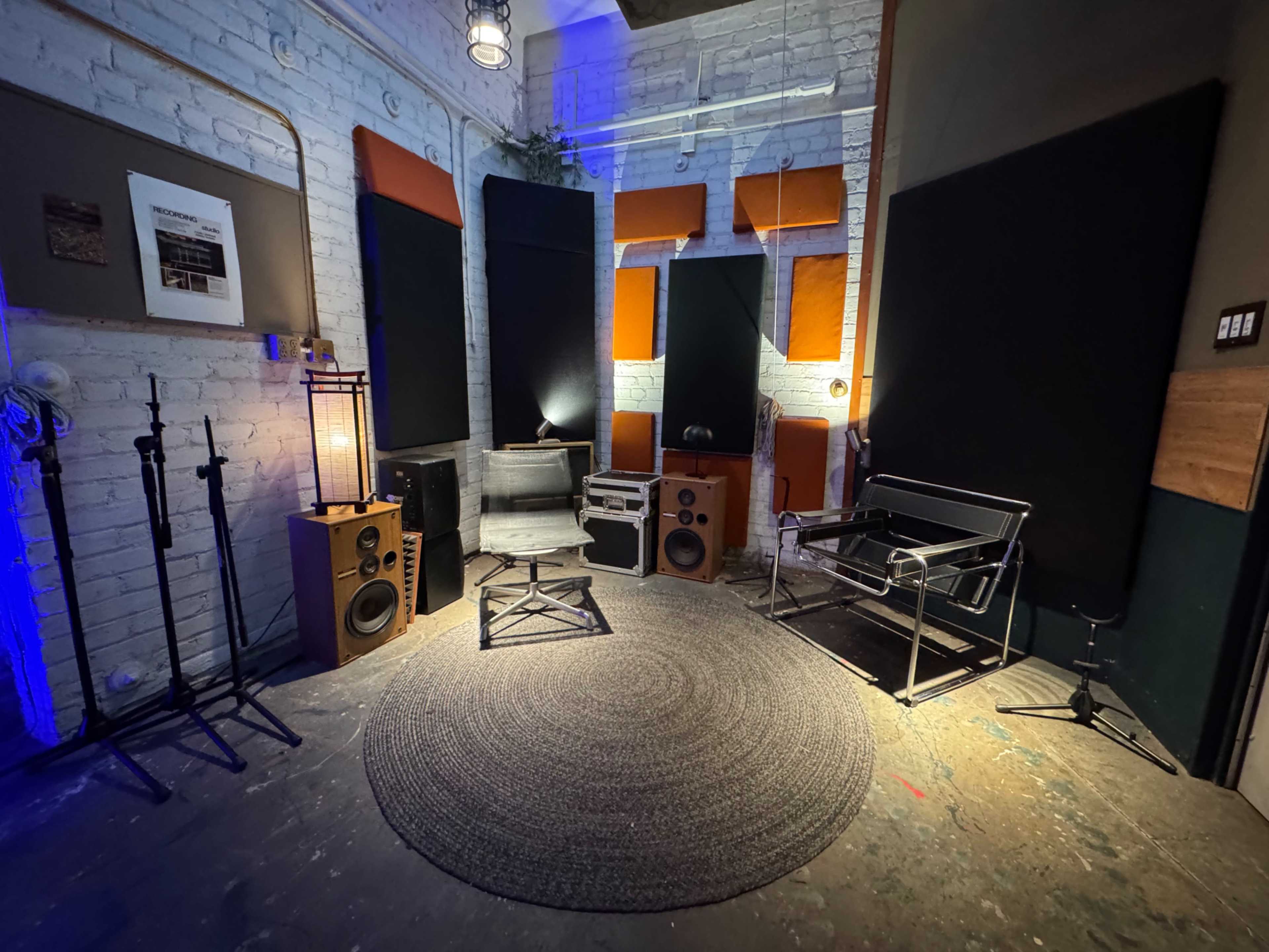 The image shows a sound studio corner featuring various audio equipment, soundproofing panels, and minimalist furniture arranged around a circular rug.