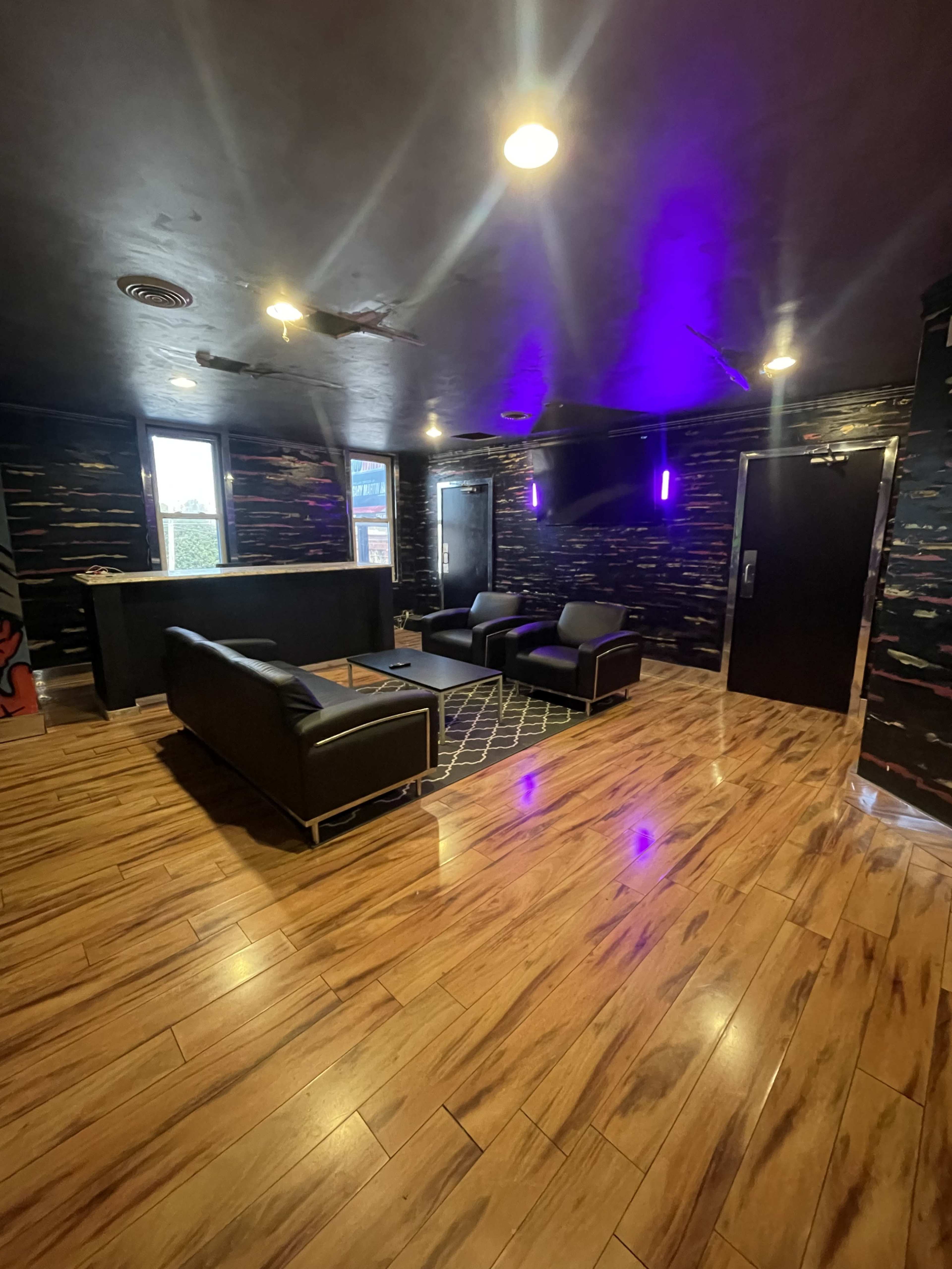 The image shows a modern lounge area with dark walls, wooden flooring, a seating arrangement of black sofas, and purple accent lights.