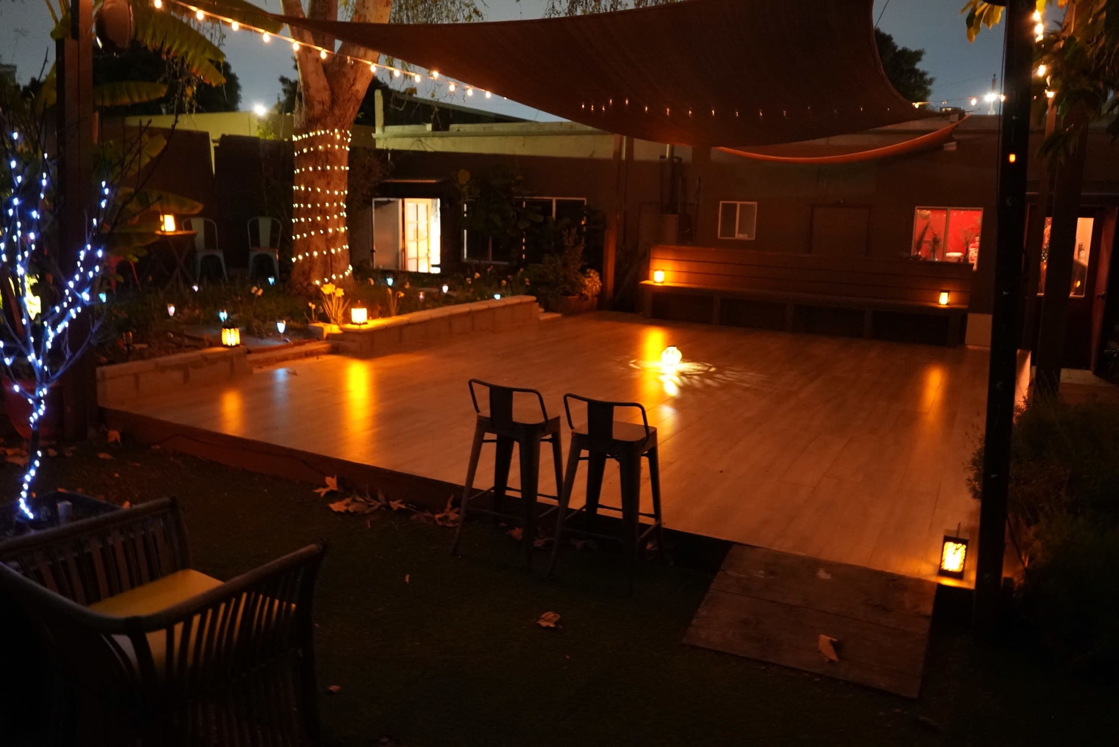 The image shows a well-lit outdoor space with a wooden platform, string lights draping overhead, and seating areas surrounded by trees and decorative lanterns.