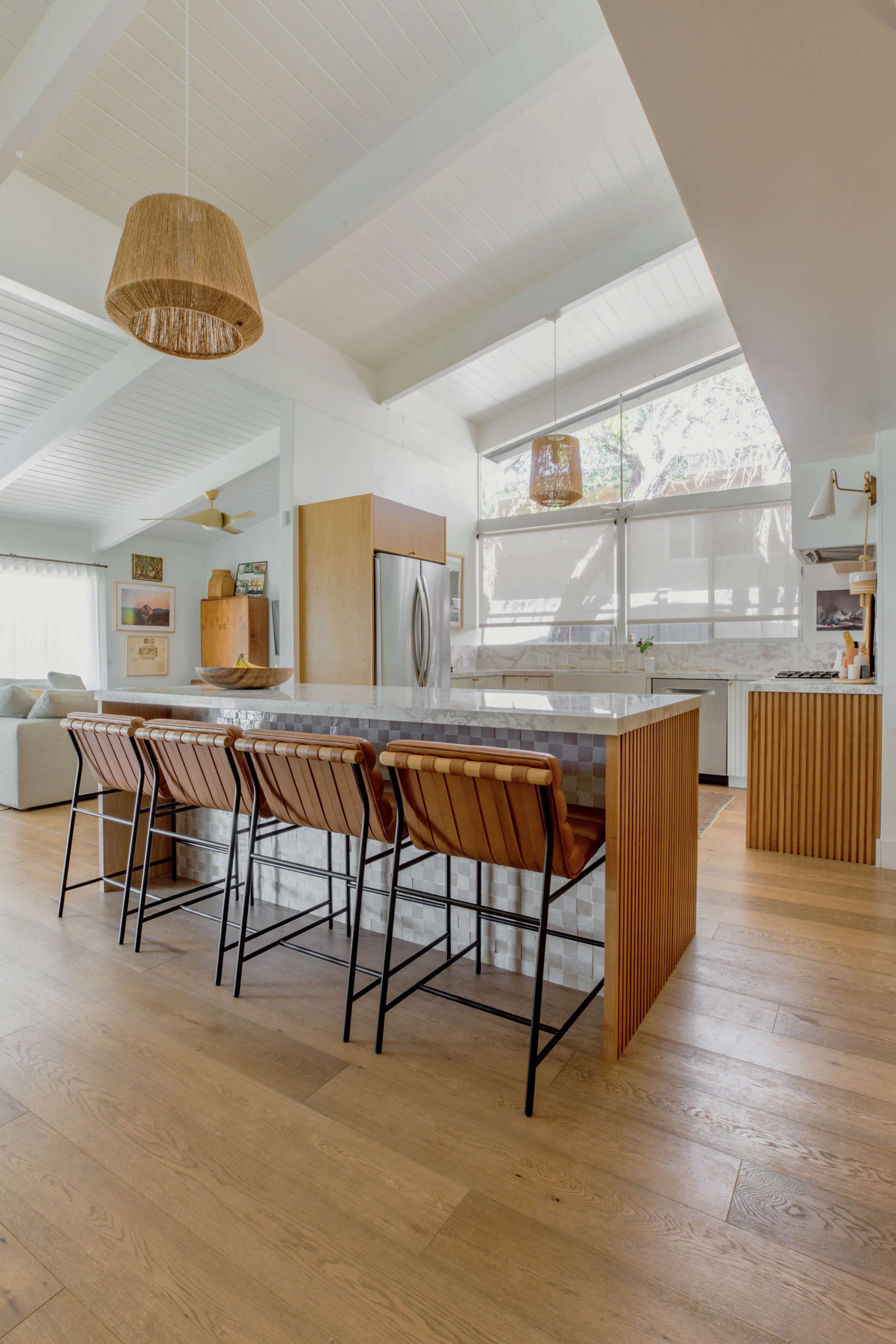 A modern kitchen features a marble island with four metallic bar stools and large windows that provide natural light.
