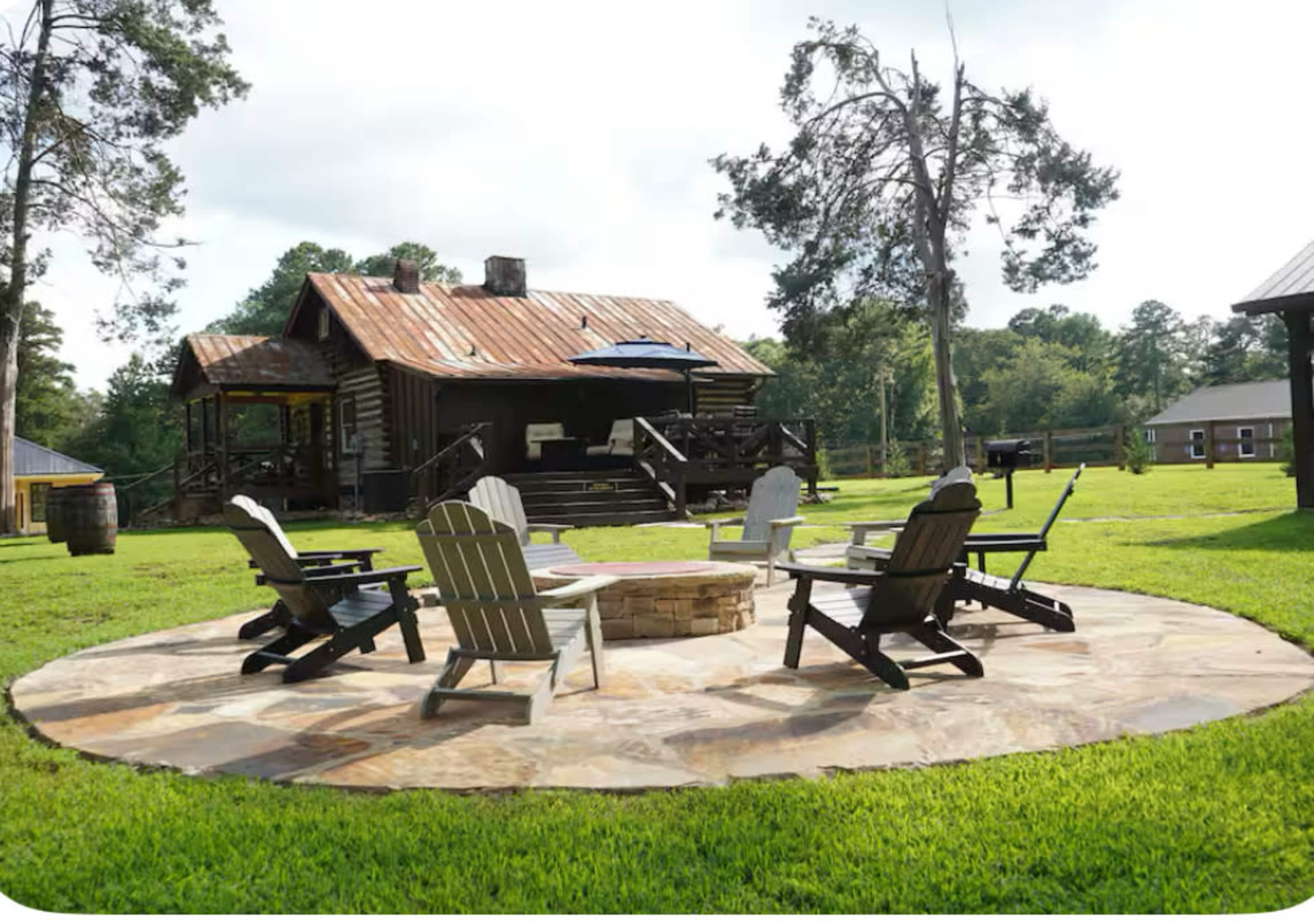 A circular stone patio is surrounded by five wooden Adirondack chairs, positioned around a fire pit in a grassy yard with a log cabin and trees in the background.
