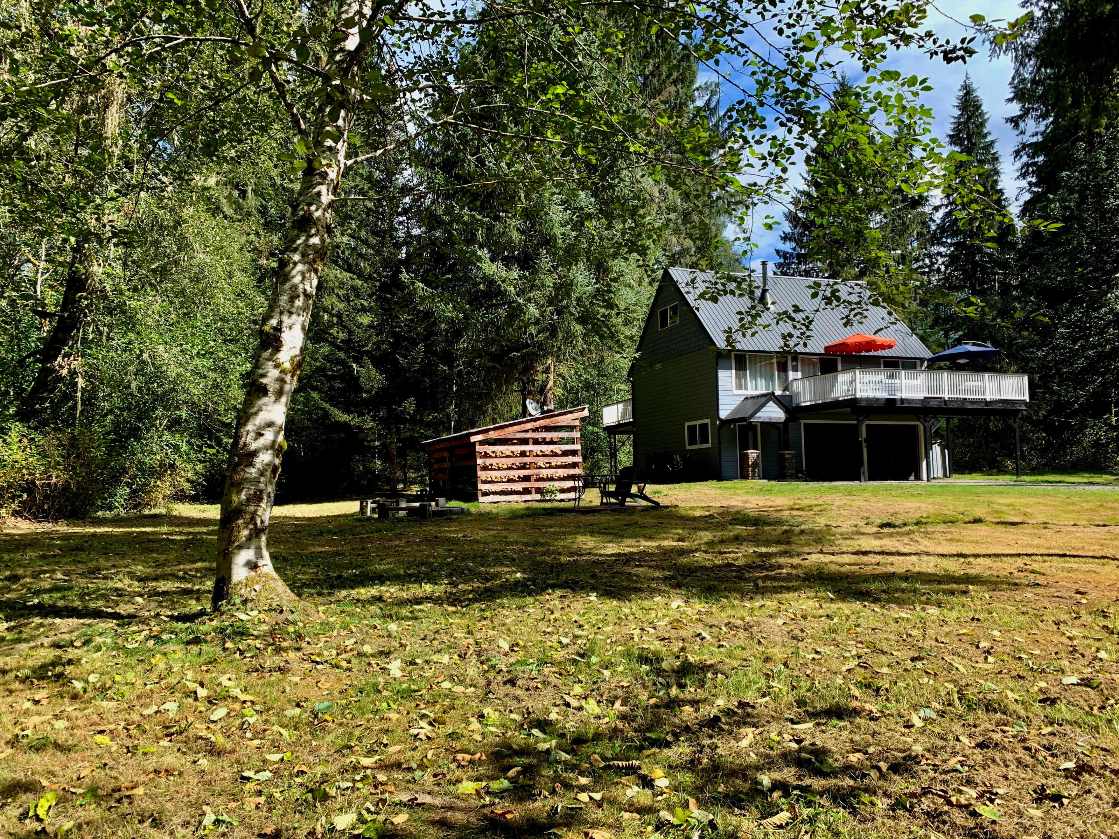 A green two-story house with a deck and an umbrella sits surrounded by trees in a grassy clearing.